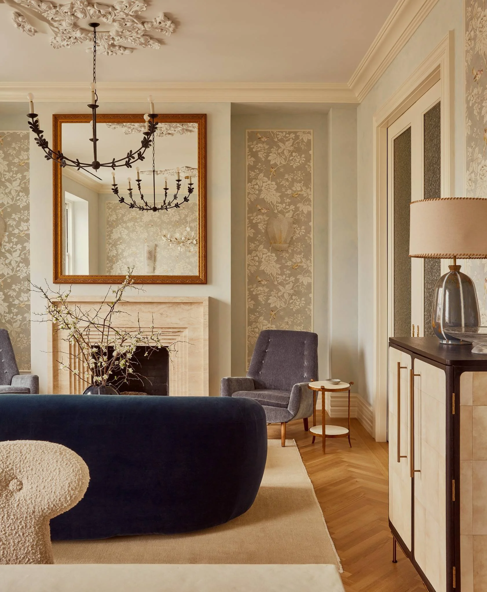 Elegant living room with a fireplace, a large mirror above it, gray armchairs, a dark blue sofa, wooden flooring, a cream-colored rug, floral wallpaper, a side table, and a chandelier hanging from an ornate ceiling medallion.
