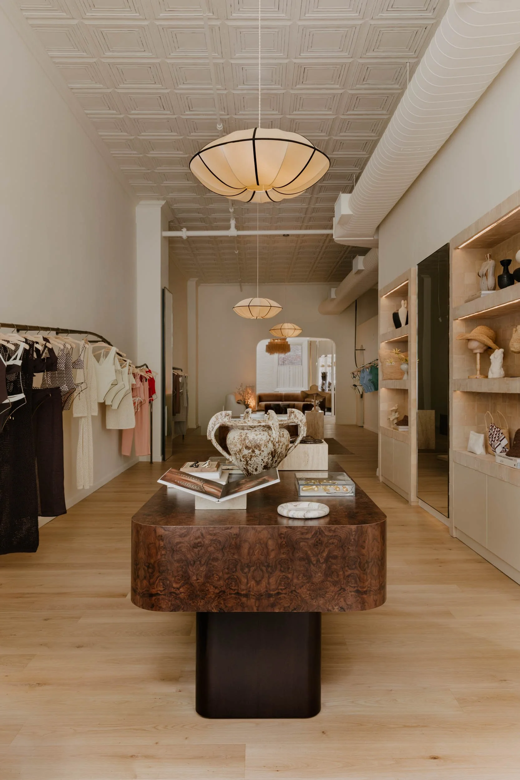 Interior of a boutique store with clothing racks on the left, shelves with decorative items on the right, and a central wooden table displaying a large ancient-style ceramic urn, magazines, and jewelry.