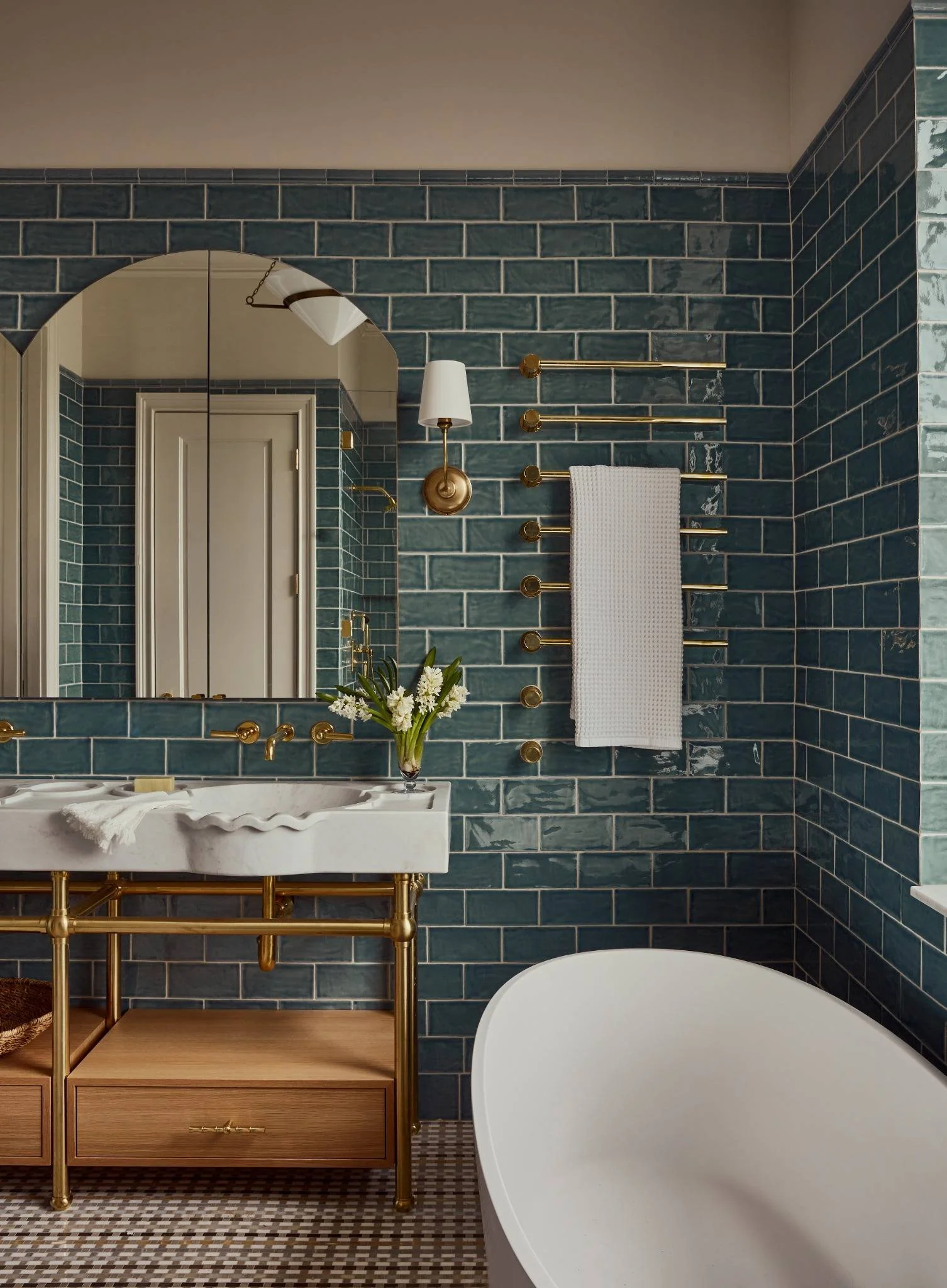 Bathroom with dark green tiled walls, a white freestanding bathtub, a marble sink with gold fixtures, a wooden vanity, a mirror, a wall-mounted towel rack, and a wall sconce with a white shade.
