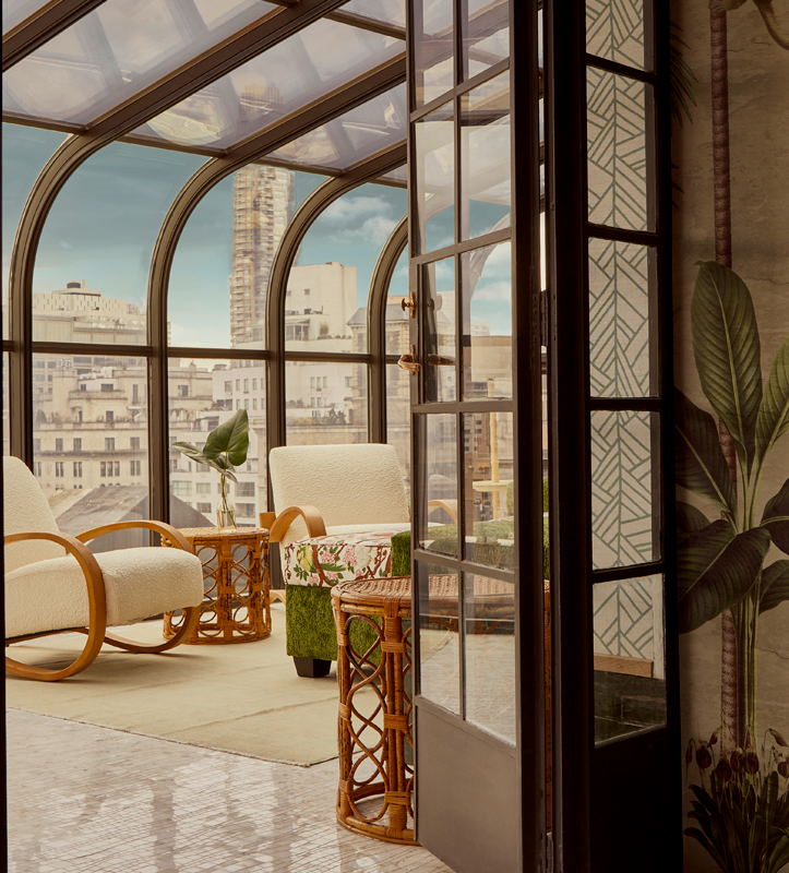 Cozy sunlit indoor space with rattan furniture, including cream armchairs, a floral-patterned armchair, and small tables, surrounded by large glass windows with an urban cityscape view.