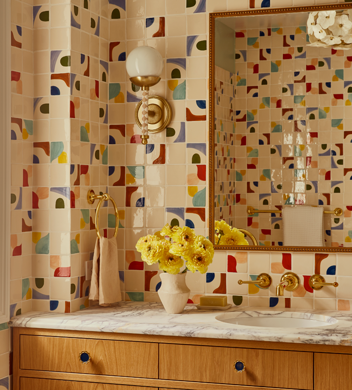 A bathroom with colorful abstract tile walls, a mirror, a marble countertop, a vase with yellow flowers, and gold fixtures.