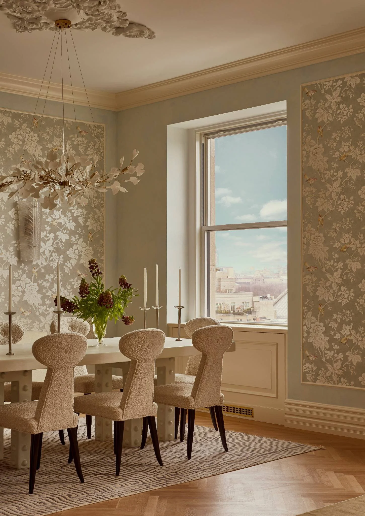 A dining room with a large window showing a cityscape and partly cloudy sky, a white table with a floral arrangement and four beige upholstered chairs, patterned wallpaper, a decorative ceiling fixture, and a patterned rug.