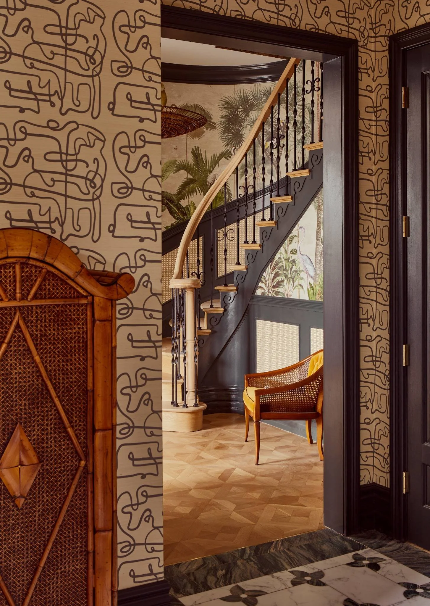 Interior view of a home with staircase, dark wood paneling, vintage wallpaper with tropical plants and herons, and a yellow upholstered chair.