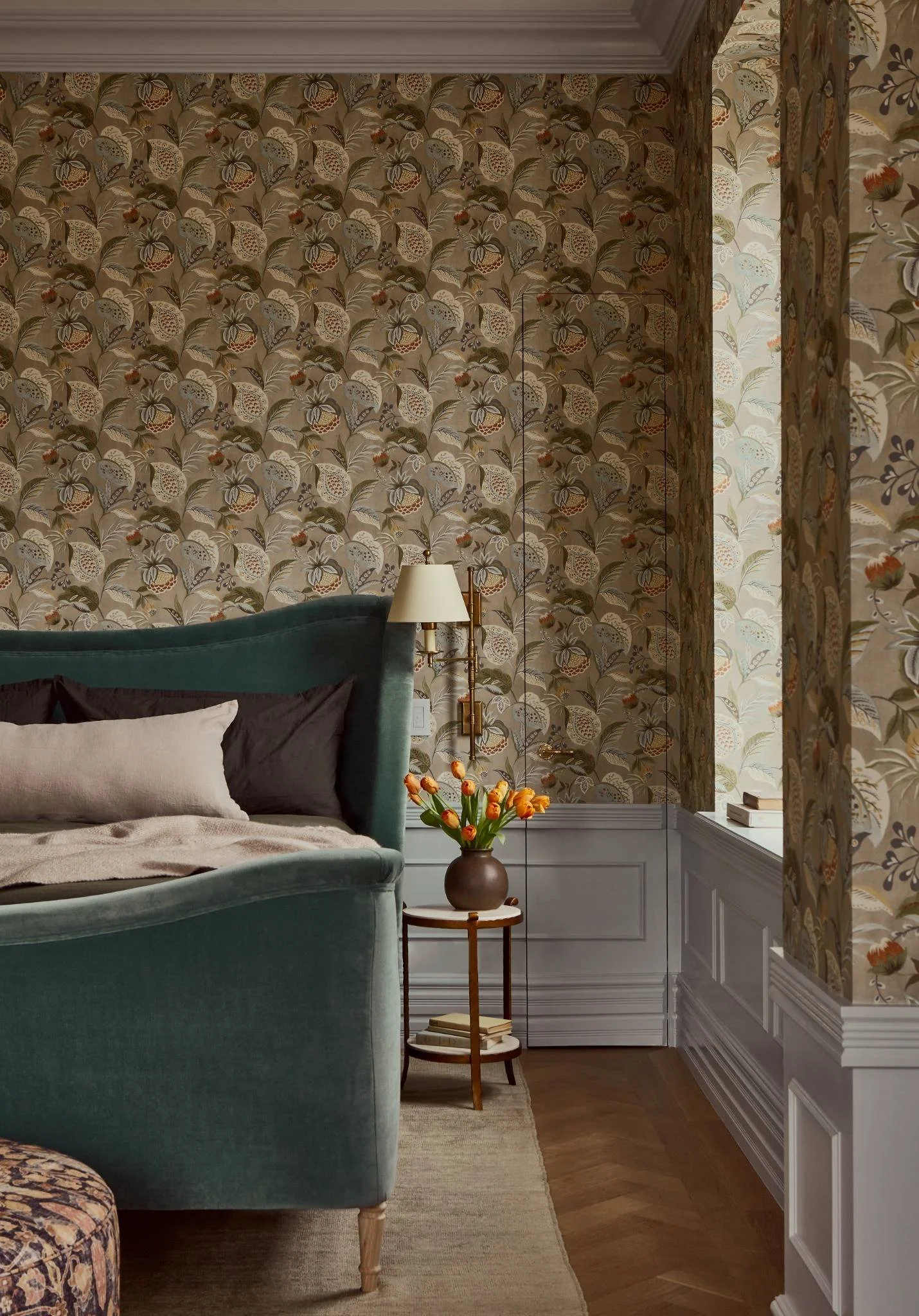 A cozy bedroom corner with floral wallpaper, a green upholstered headboard, a white pillow, a side table with a vase of orange tulips, a wall-mounted lamp, a window with books, and wood flooring.