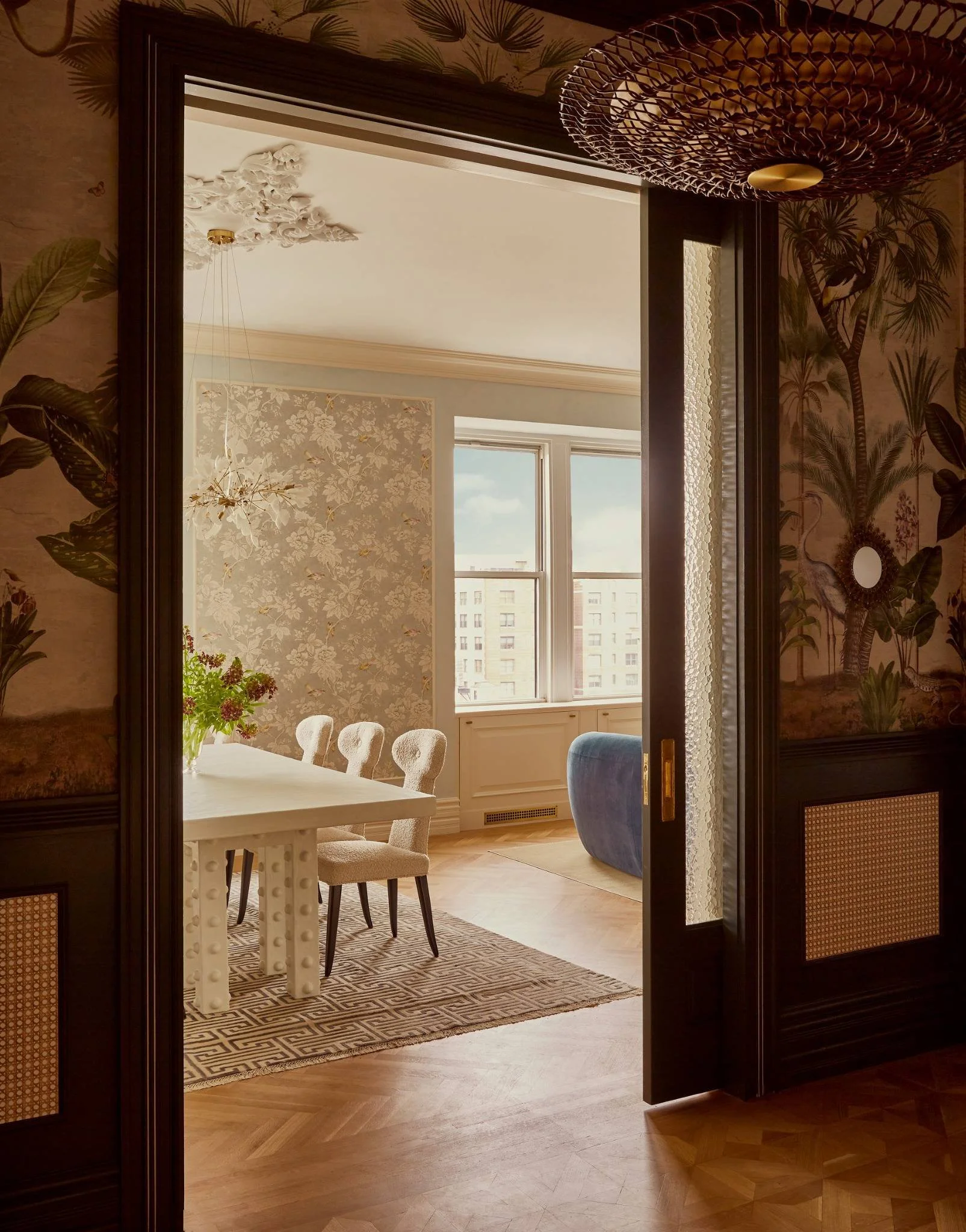 View of a bright dining room with a white table, four textured beige chairs, a vase of flowers, and a large window showing a cityscape.