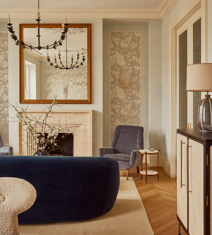Elegant living room with a large mirror above a fireplace, chandelier, armchair, sofa, side table, and decorative wallpaper.