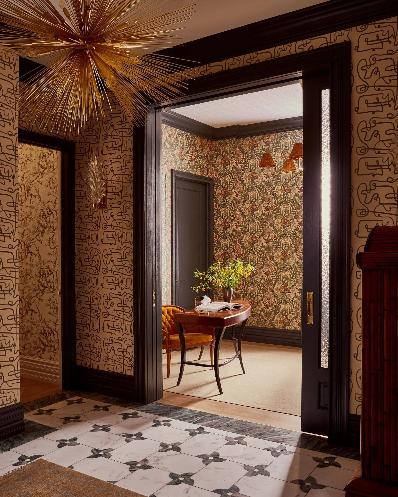 A view through a doorway into a room with patterned wallpaper, a small wooden table, a chair with a tufted seat, a vase with green foliage, and wall-mounted lamps.