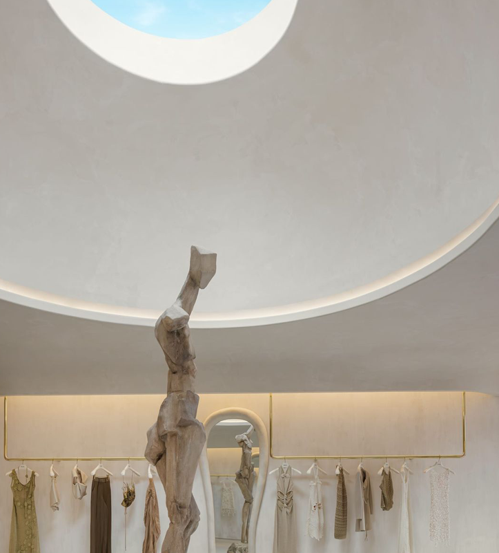 Interior of a boutique store with a wooden sculpture in the foreground, a gold clothing rack with dresses and blouses, and a large circular ceiling window letting in natural light.