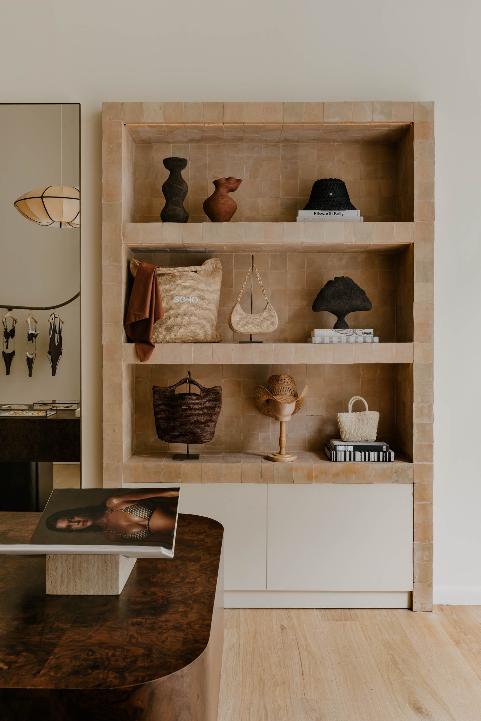 A beige tiled shelving unit with decorative items like vases, books, and handbags. A small table with a fashion magazine is in front of the shelving unit. To the left, part of a mirror and a hanging jewelry display are visible.