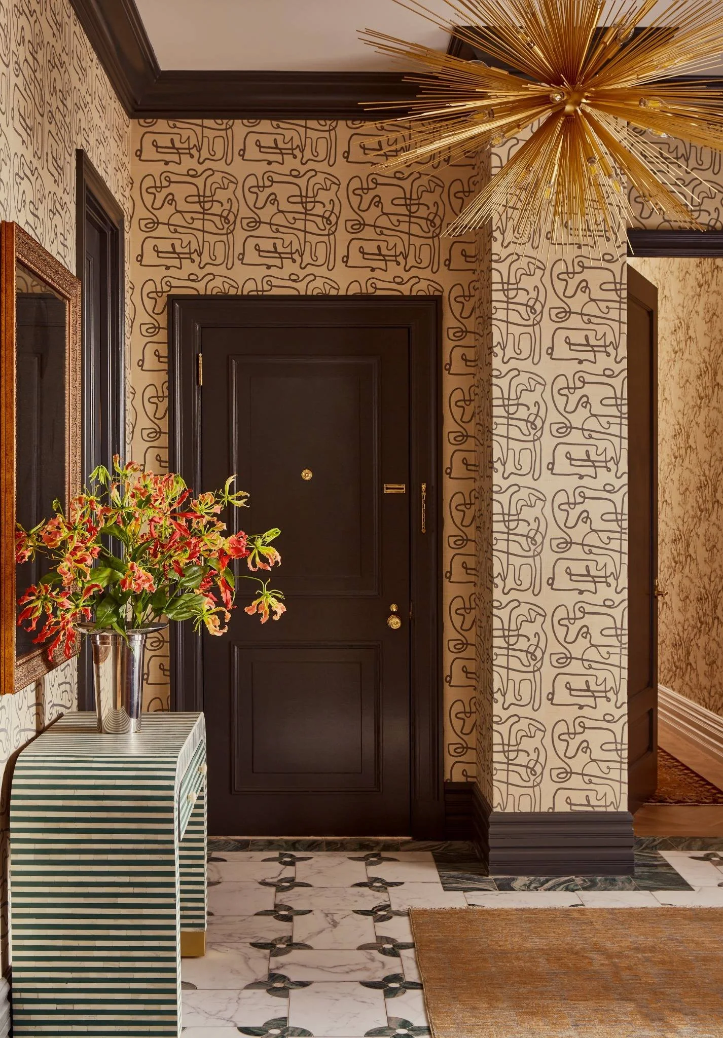 Entryway with black door, patterned wallpaper, marble floor, gold chandelier, mirror, and a striped table with a flower arrangement.