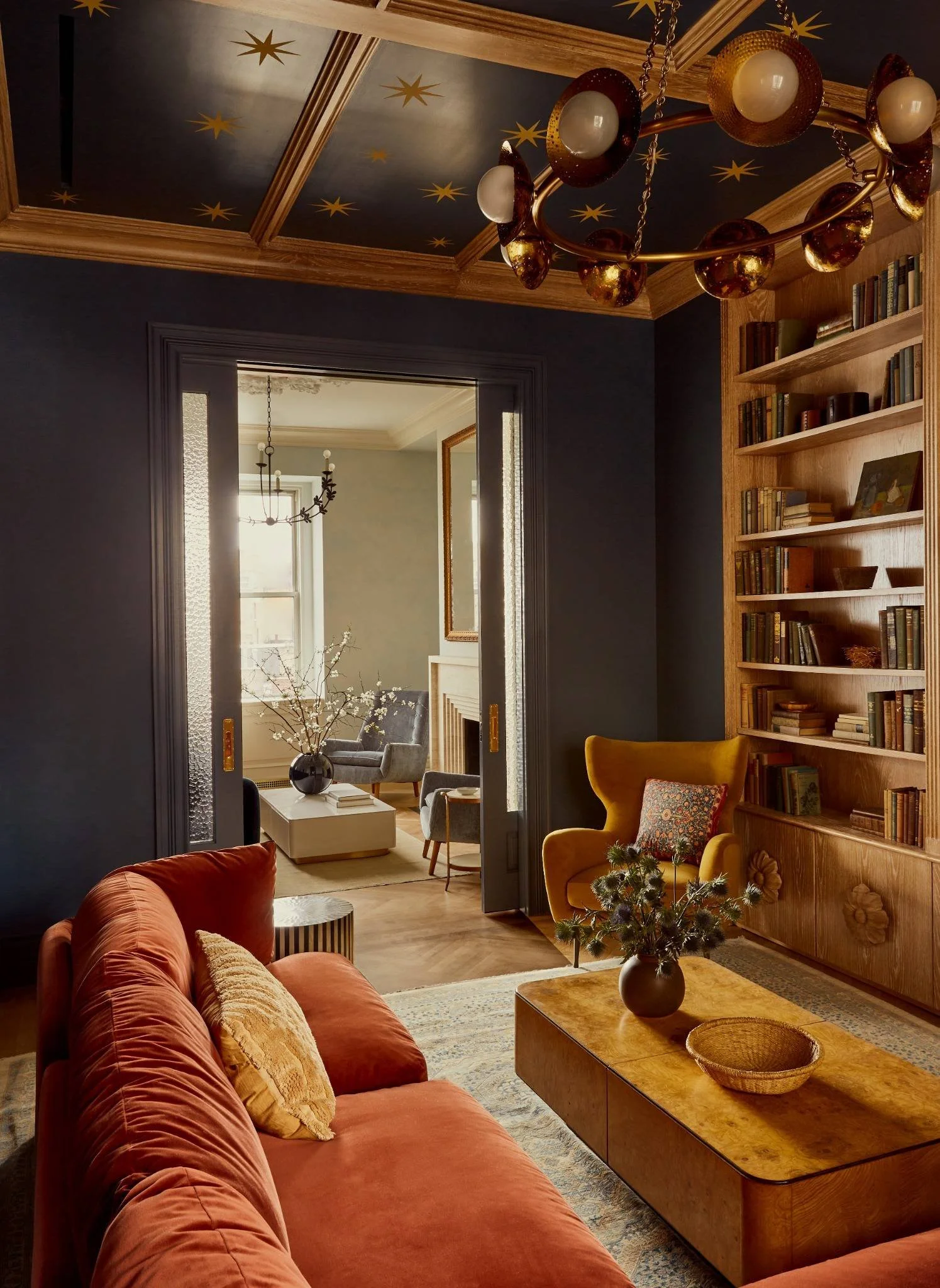 Living room with a burnt orange sofa, yellow armchair, wooden bookshelf, coffee table with a vase of flowers, and a dark ceiling with gold star decorations. Through open doorway, another sitting area with gray chairs, a white coffee table, and a chandelier is visible.