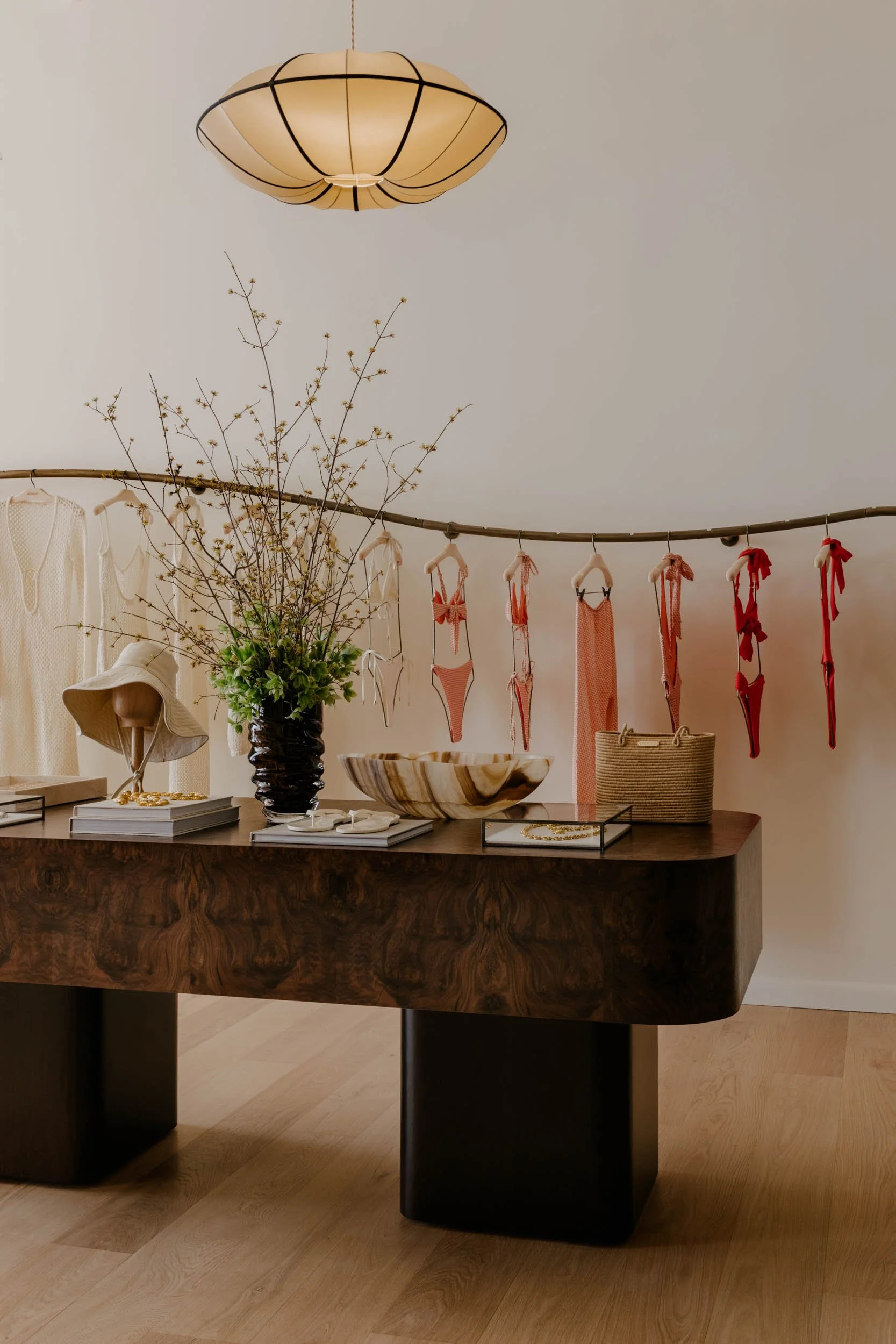 A retail display area with a wooden table showing various fashion and home accessories, including a large hat, jewelry, vases, and clothing on a rack. A branch with flowers decorates the background, and a ceiling light fixture is visible.