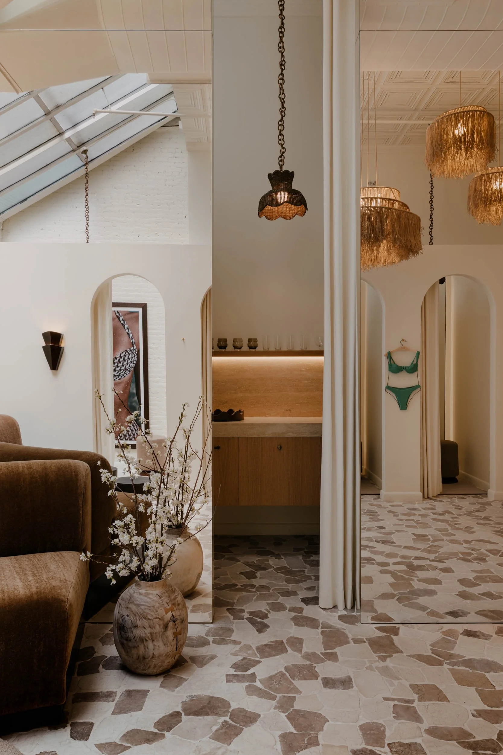 Interior with a beige armchair, a ceramic floor vase with white flowers, a mirror, pendant lights with fringed shades, and arched doorways leading to other rooms.