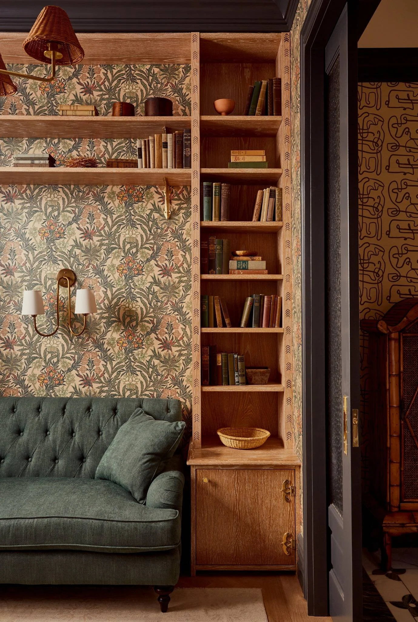 A cozy living room corner featuring a green tufted sofa, a wooden bookshelf filled with books, decorative objects, and a wallpapered wall with floral and botanical patterns. There is a wall-mounted lamp with two white lampshades on the wallpapered wall.