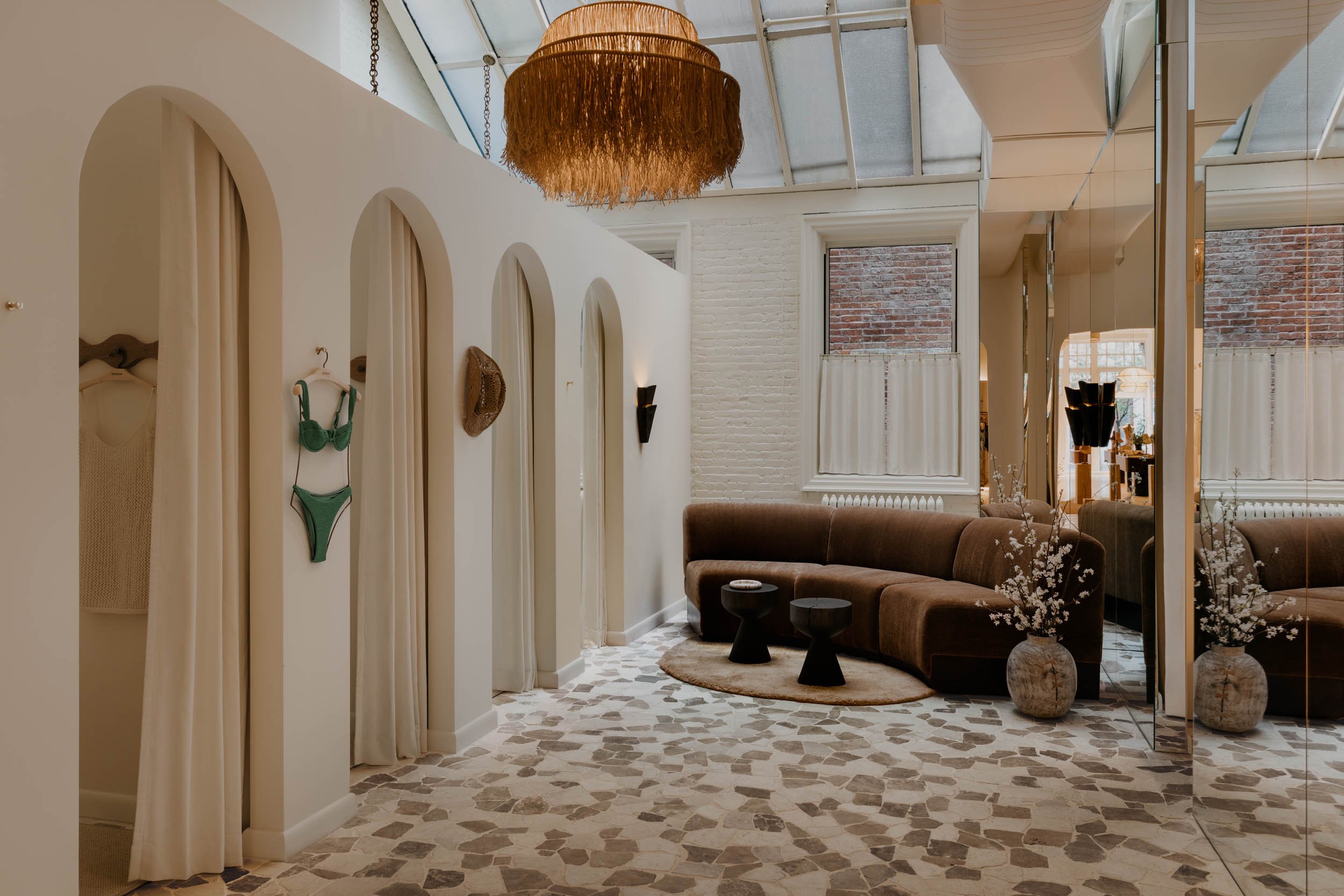 Interior view of a cozy, modern lounge area with a brown sectional sofa, small black tables, and decorative plants. The space features a ceiling with large glass panels, a trendy woven light fixture, and a wall with arched niches displaying clothing and accessories. The floor has a mosaic-style pattern, and windows with white curtains let in natural light.