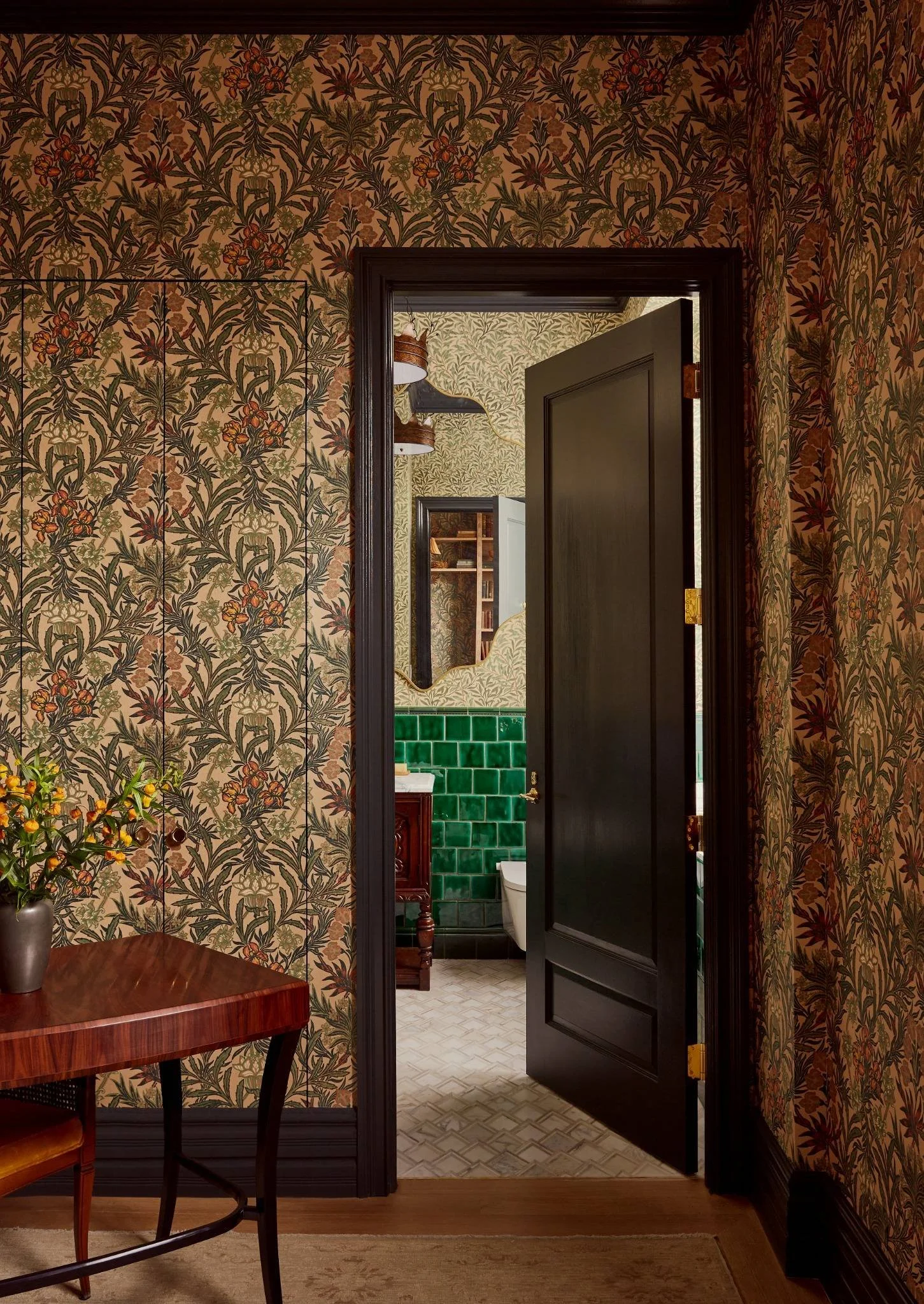 A view into a bathroom through an open dark door. The bathroom has green tiled walls, a white toilet, a mirror, and a wooden vanity. The room outside the bathroom features floral wallpaper in earth tones, a wooden table with a vase of flowers, and patterned wallpaper in the background.