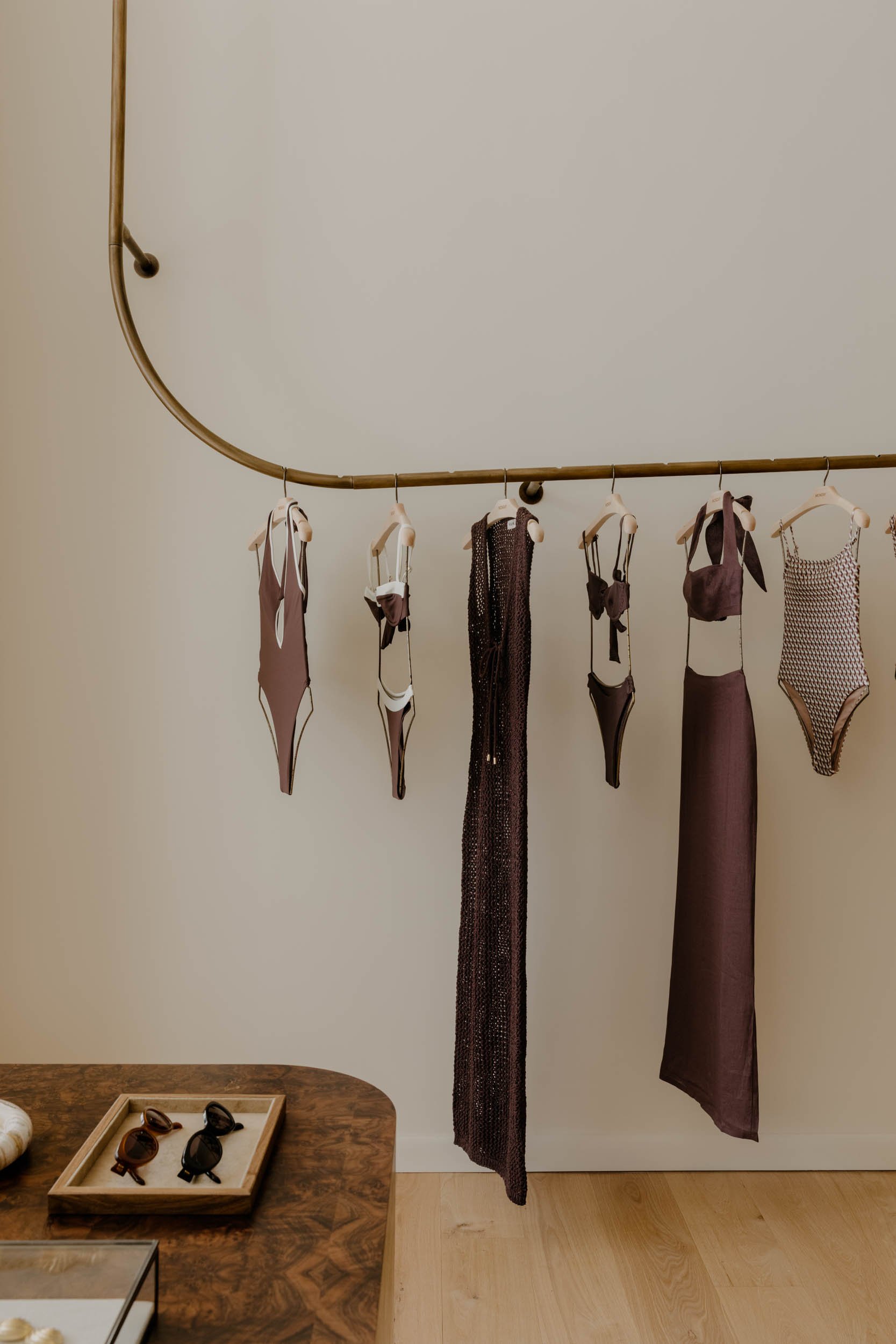 Display of clothing and accessories, including various bras and a dress on a hanging rod, with sunglasses on a wooden table below.