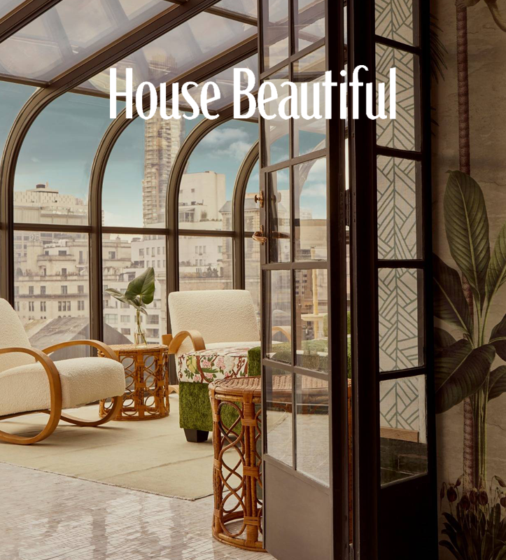 Bright sunlit indoor seating area with large arched windows showing city buildings outside. Includes white armchairs, a small wooden side table with a potted plant, and patterned cushions.