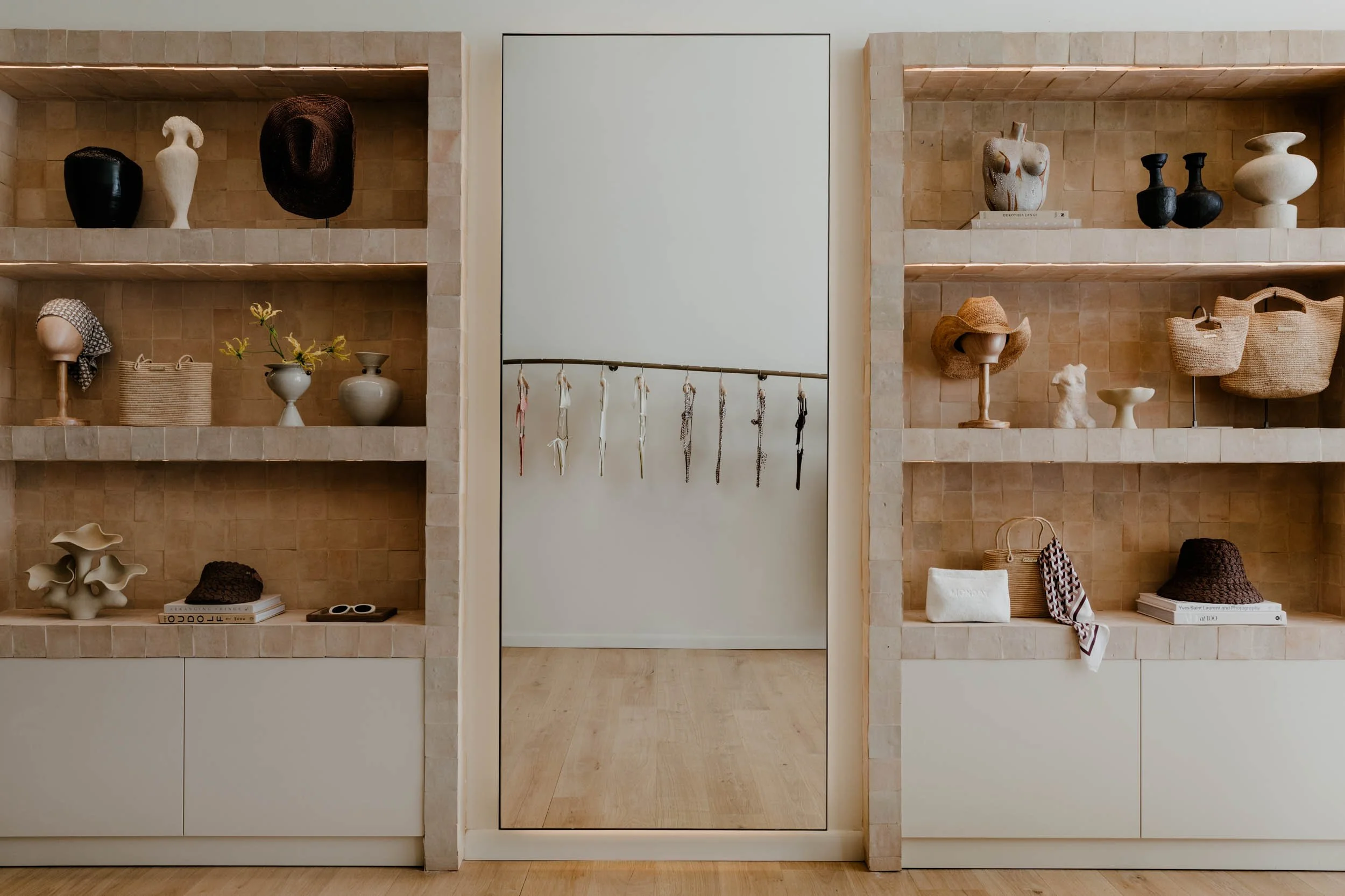 Open shelving unit with decorative ceramic vases, hats, woven bags, and books, against a beige tiled wall with a mirror in the center.