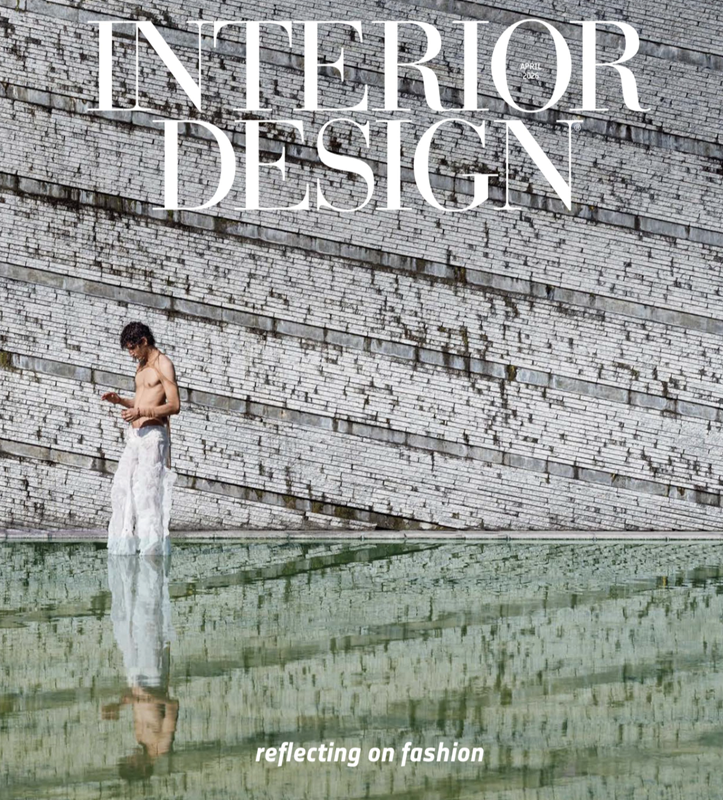 A woman in a white skirt stands near a reflective water feature in front of a curved brick wall, looking at her phone. The wall has large text that reads 'INTERIOR DESIGN' and smaller text that says 'April 2023' and 'reflecting on fashion'.