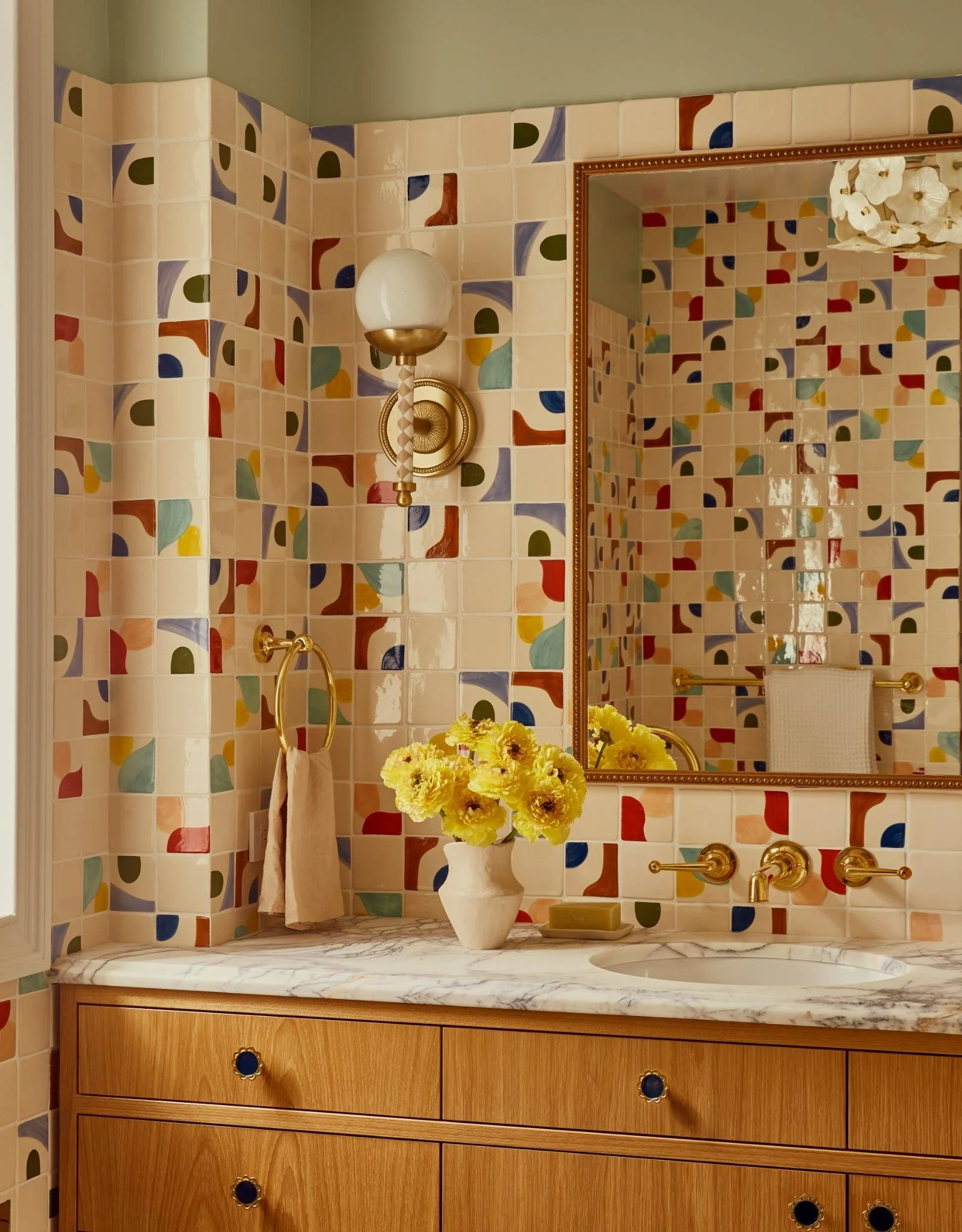 A bathroom with colorful geometric patterned tiles on the walls, a marble countertop with a vase of yellow flowers, and a large mirror reflecting the tiled wall. There is a wall-mounted light fixture with a globe-shaped bulb and gold accents, a gold towel ring with a beige towel, and gold faucet fixtures.