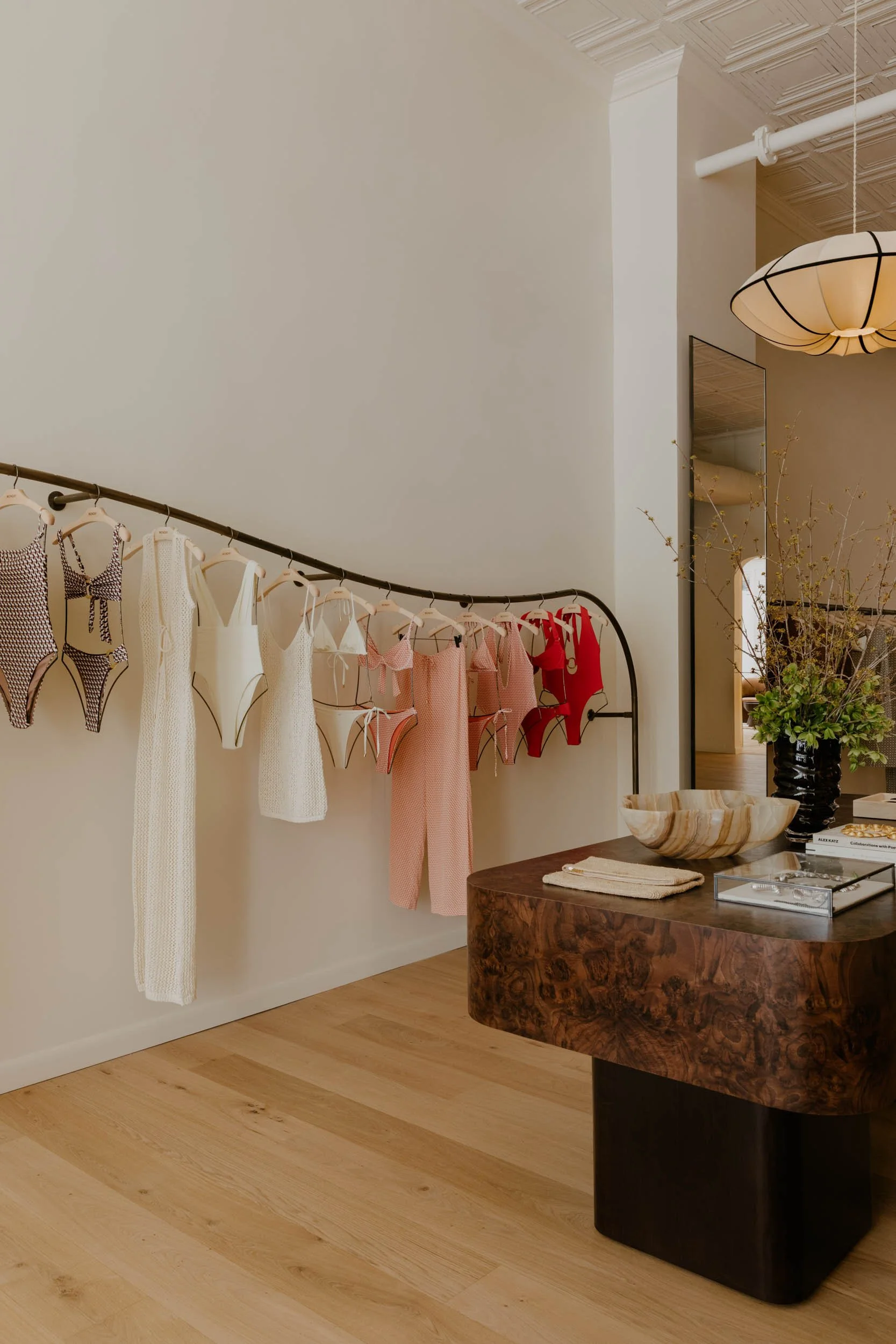 A retail store display of various swimsuits hanging on a curved black metal rack, with a wooden table holding a bowl and other items nearby.