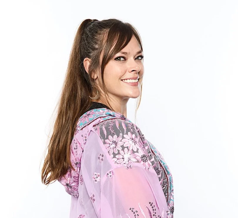 Molly Moon Fox, a Psychic with long light brown hair in a high ponytail, smiling and looking over her shoulder, wearing a sheer purple floral embroidered jacket.