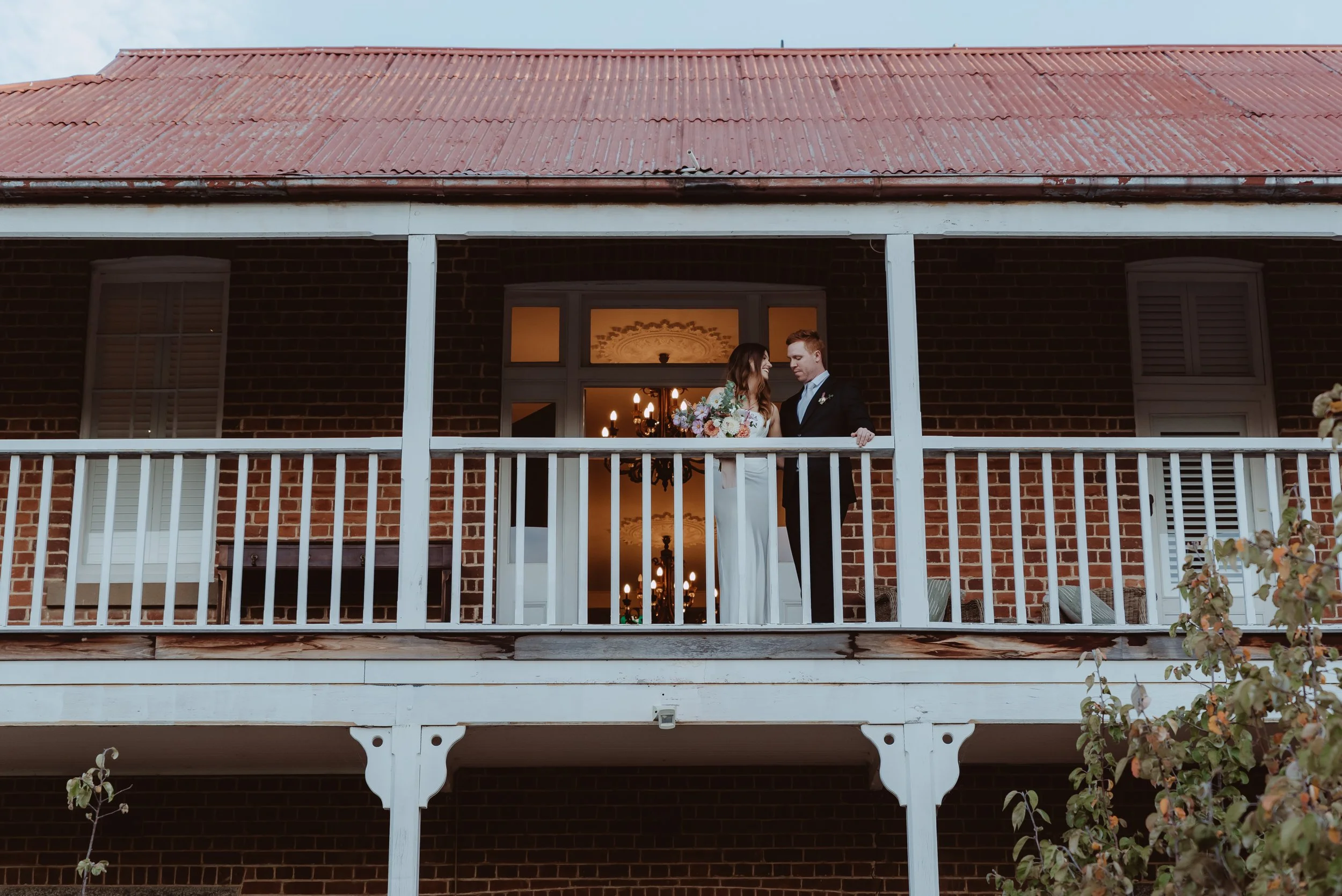A bride and groom standing on a balcony of The Doncaster in Braidwood, NSW. Inside and chandelier with lit bulbs visible through the window behind them.