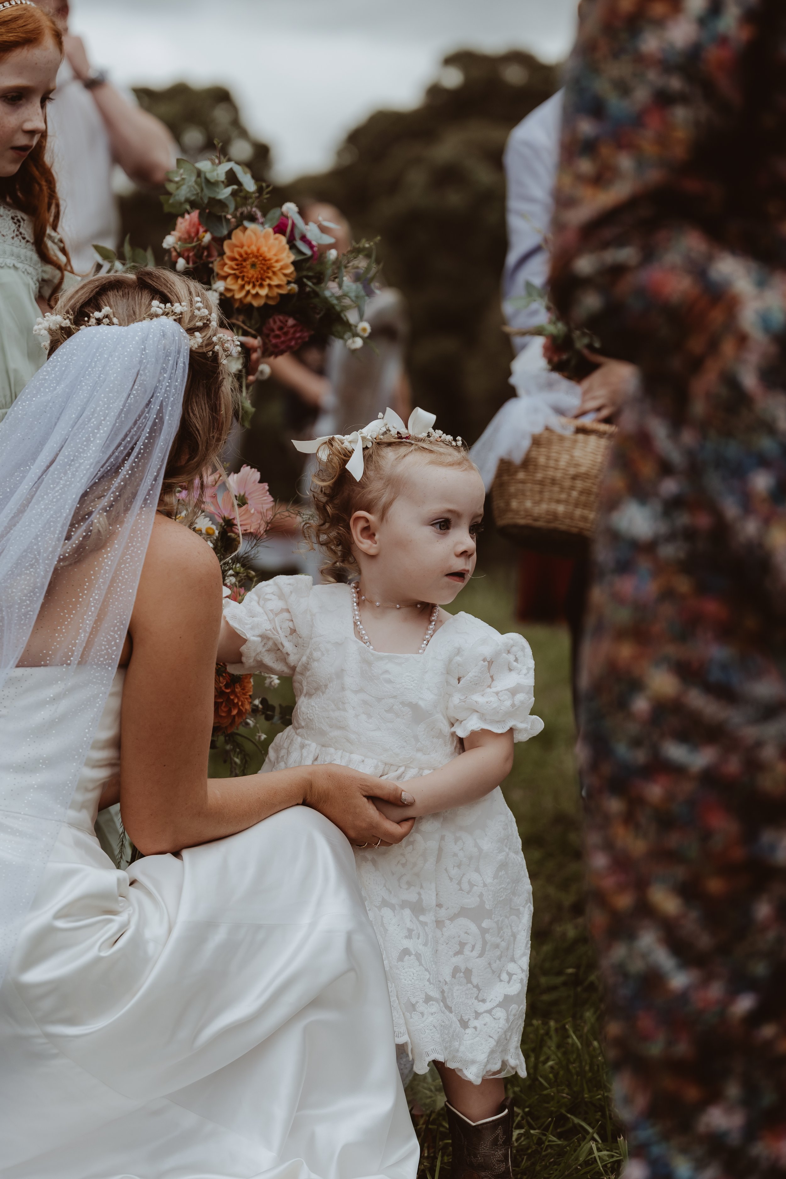 Braidwood Wedding Photographer. A young girl dressed in a white lace dress with a pearl necklace and a floral headband with a bow, participating in an outdoor ceremony, possibly a wedding, surrounded by women and children holding bouquets of flowers.