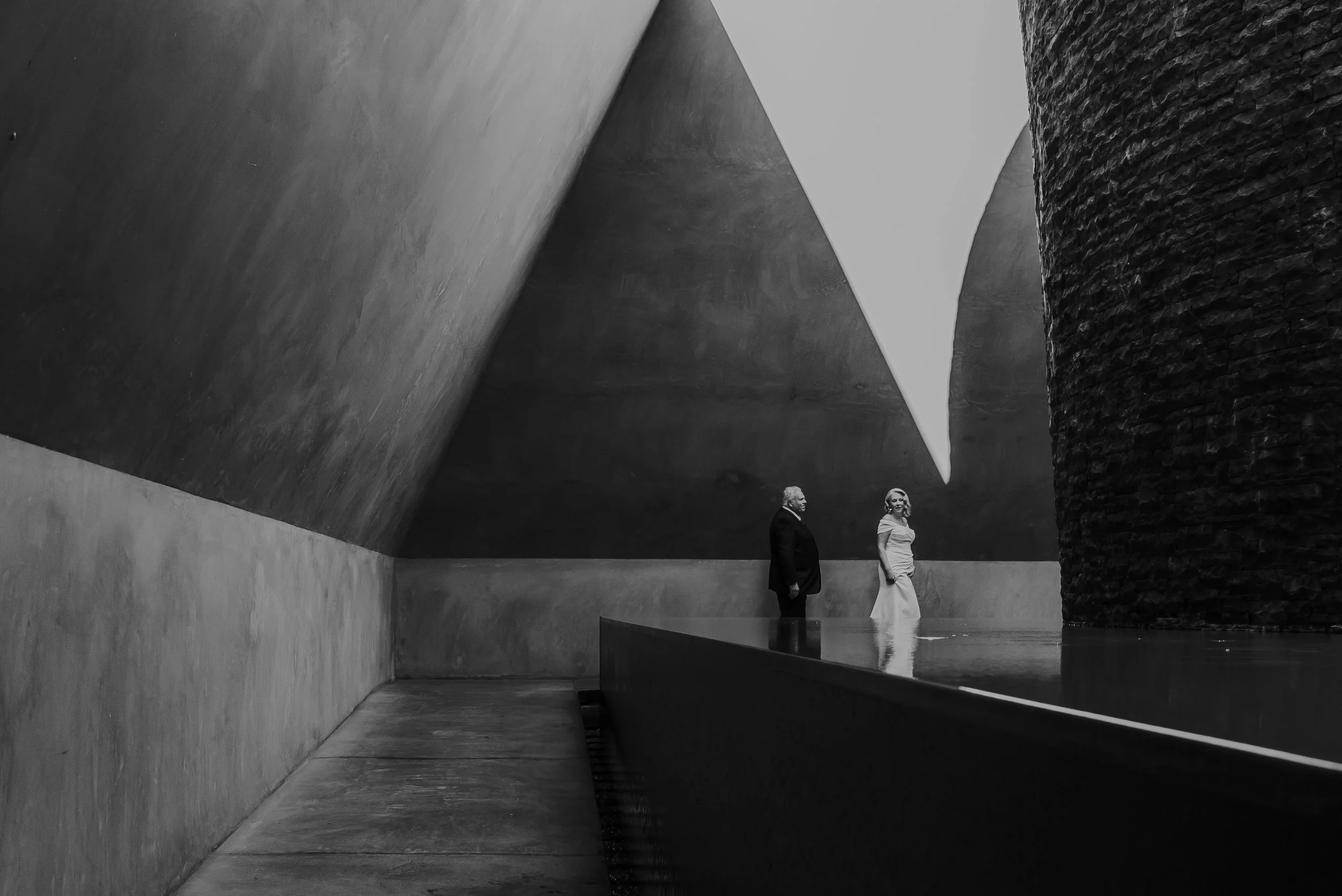 Braidwood Wedding Photographer. Black-and-white photo of a modern, abstract architectural interior with large angular concrete walls and a textured brick column. Two elegantly dressed people, a man in a dark suit and a woman in a white dress, stand o