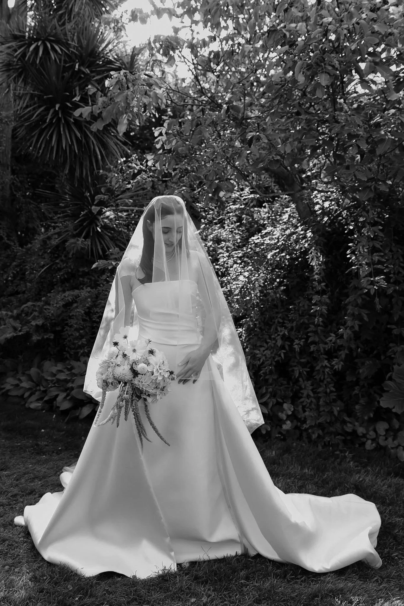 Braidwood Wedding Photographer. Black and white photo of a bride in a wedding dress, holding a bouquet of flowers, standing outdoors with trees and foliage in the background.