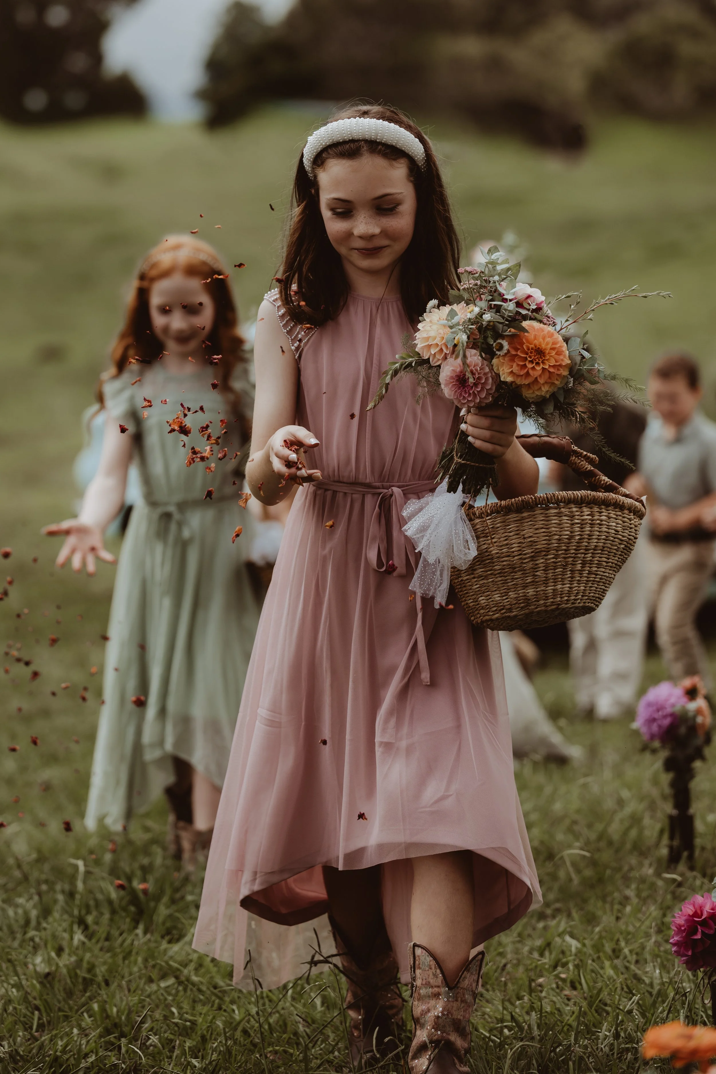 Braidwood Wedding Photographer. A young girl in a pink dress holding a bouquet of flowers walking through a grassy field, with a blurred girl and boy in the background, during daylight.