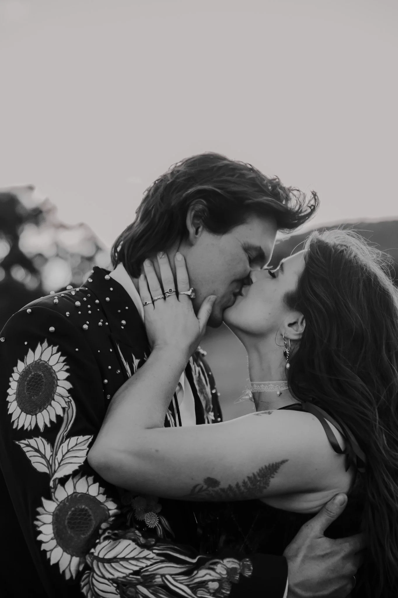 A black and white photo of a man and woman sharing a kiss at their country Canberra wedding. The man has long hair and is wearing a jacket with sunflower patterns. The woman has tattoos on her arm, is wearing earrings, and wears a choker necklace.