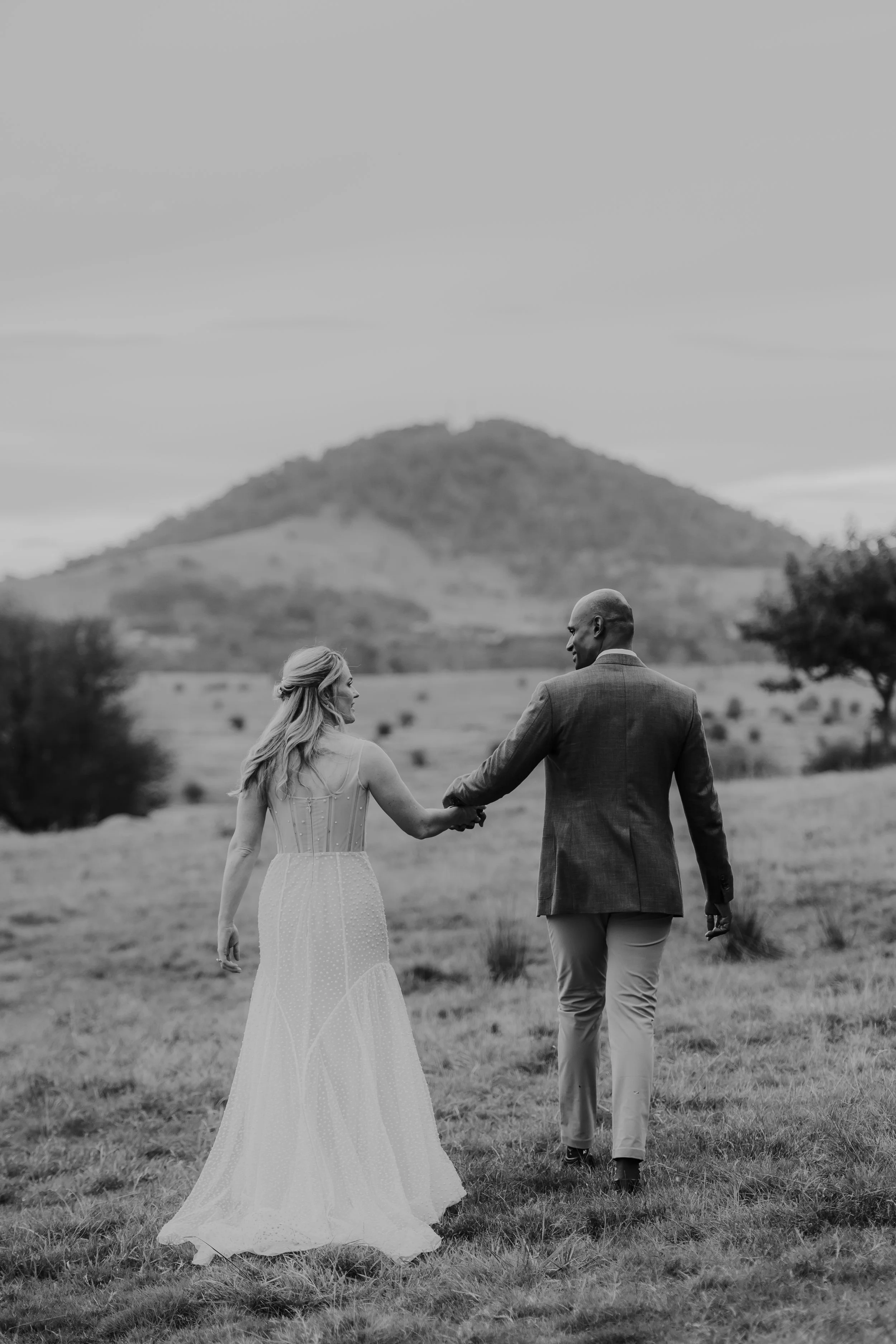 A black and white photo of a couple holding hands and walking in an open field with mount Gilamatong in the background. The woman is wearing a white dress and the man is wearing a suit. They are at their wedding in Braidwood.