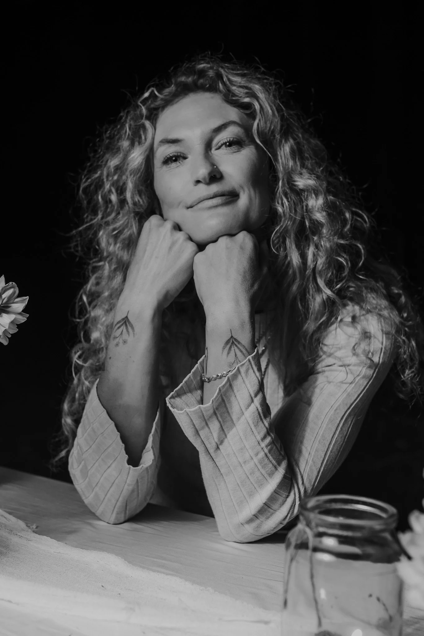 Black and white photo of a young woman with curly hair wearing a beanie, sitting outdoors on the ground covered with leaves, with a wooden fence and large tree in the background, smiling at the camera.
