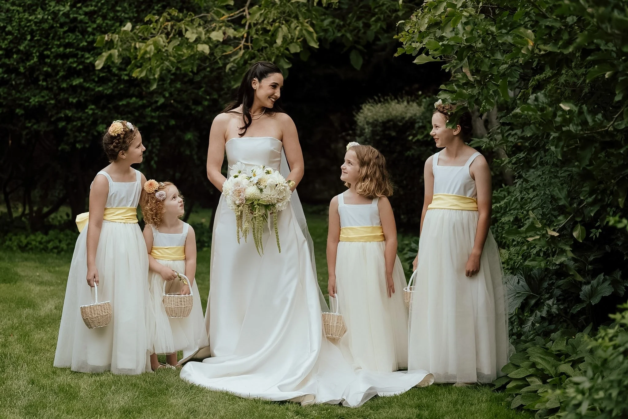 Braidwood Wedding Photographer. A woman in a white strapless wedding dress holding a bouquet of white flowers, standing outside among five young girls in white dresses with yellow sashes, green foliage in the background.