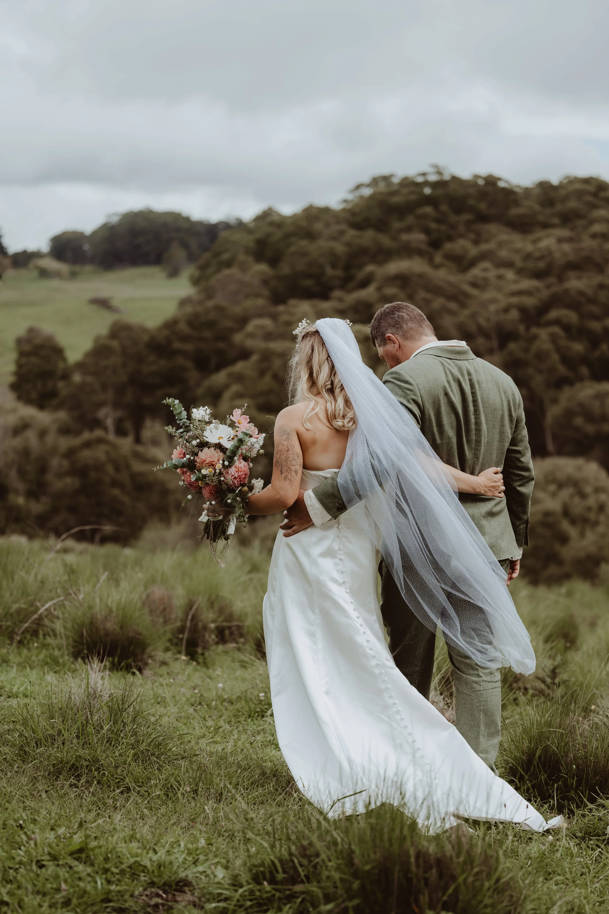 Braidwood Wedding Photographer. A bride and groom holding each other and standing on grass in a field with trees and hills in the background, during daytime, with cloudy skies.