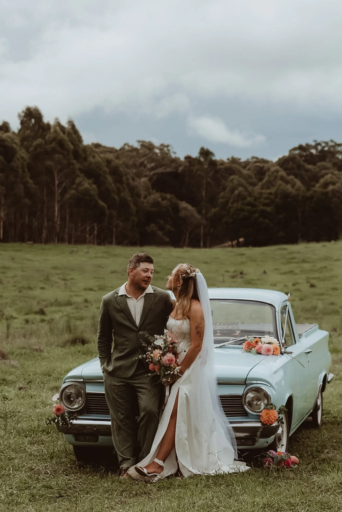 A bride and groom standing in of a vintage blue car decorated with flowers, holding a bouquet, outdoors in a green field with trees and cloudy sky in the background during a Braidwood wedding.