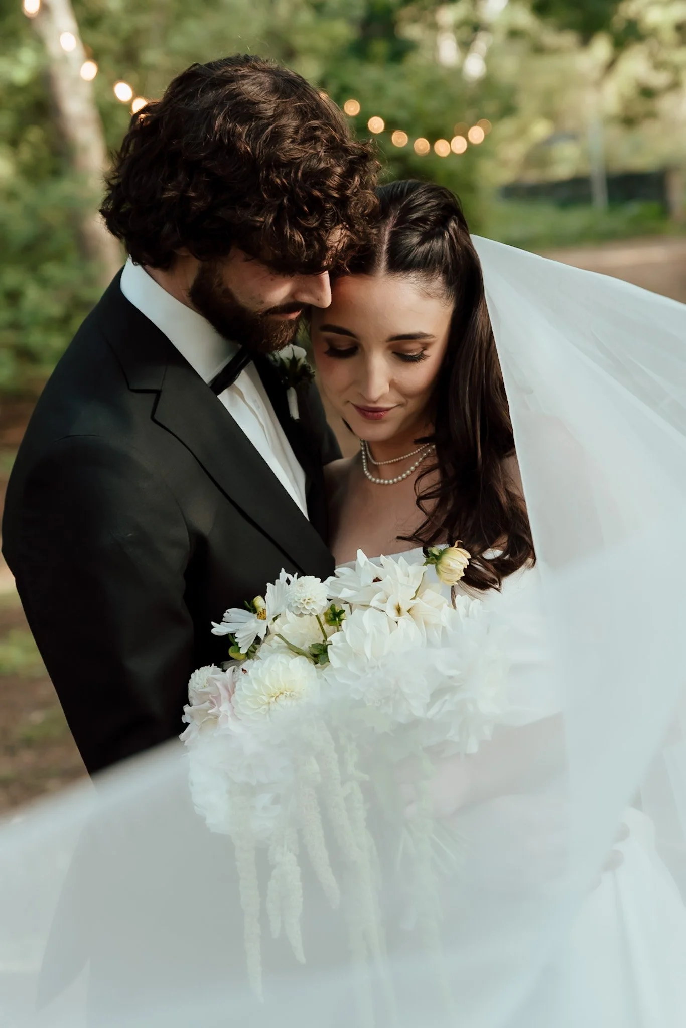 Braidwood Wedding Photographer. A bride and groom close together, holding a bouquet of white and pink flowers, outdoors with blurred green trees and string lights in the background.
