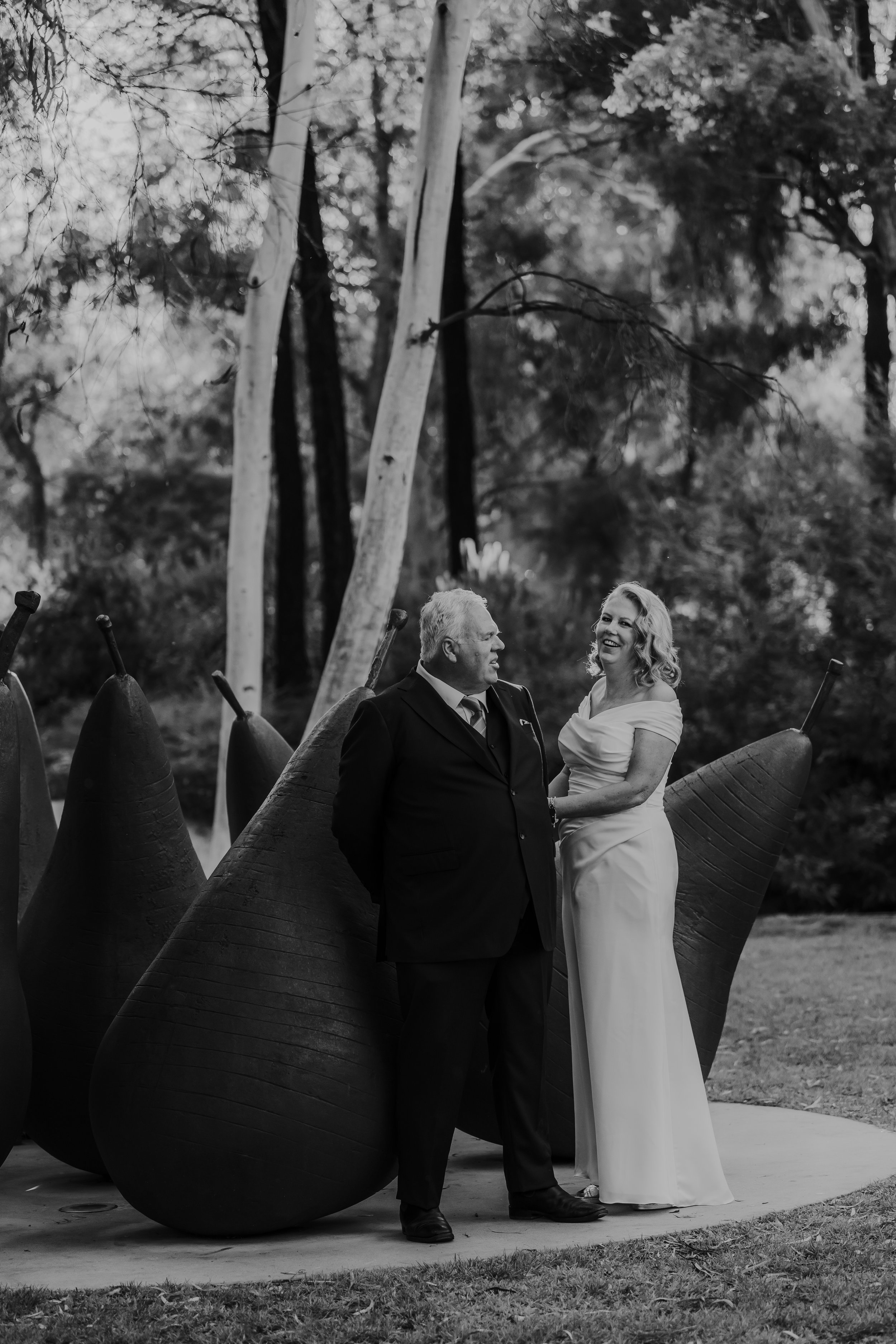 Braidwood Wedding Photographer. A black and white photograph of a man and woman dressed formally, standing closely and smiling at each other, positioned between large outdoor sculptures resembling giant berries, with trees in the background.