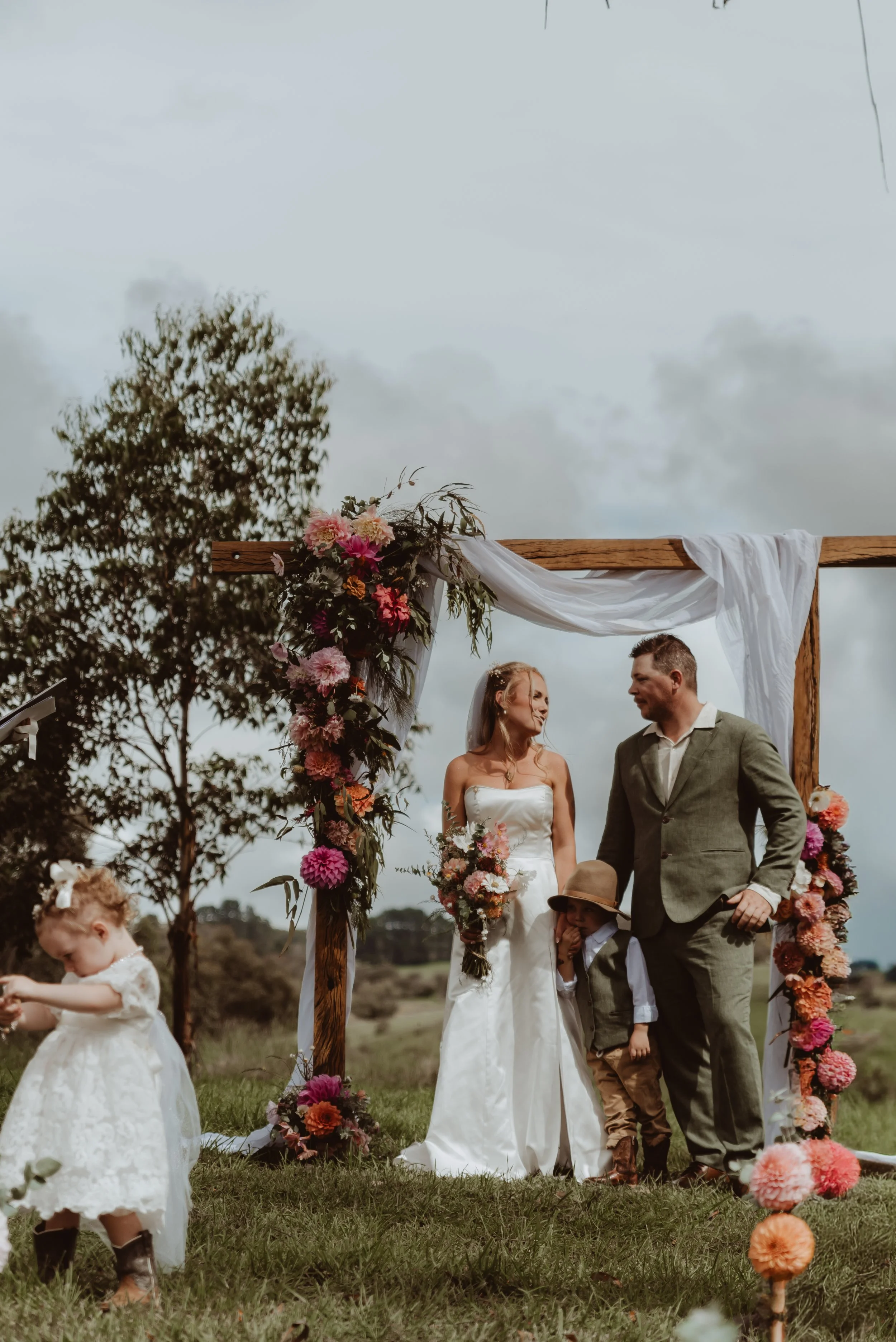 Braidwood Wedding Photographer. A wedding ceremony outdoors with a wooden arch decorated with pink and orange flowers, white drapes, and greenery. The bride in a white wedding gown and the groom in a gray suit are standing together, along with two ch
