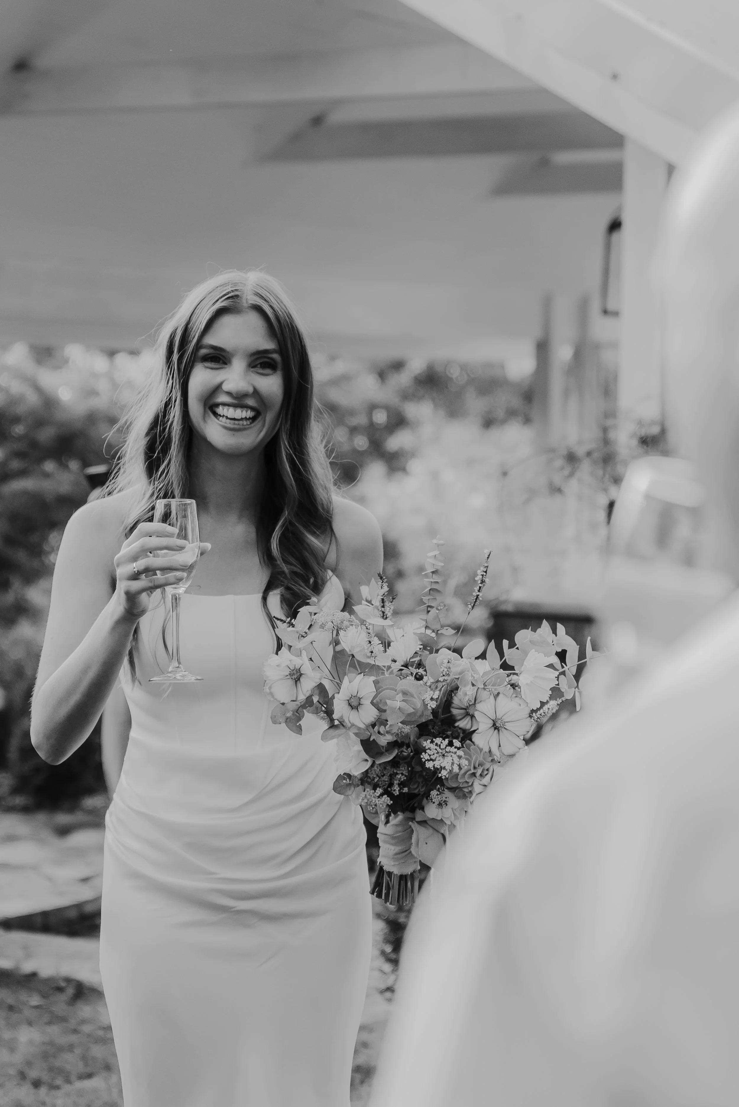 Braidwood Wedding Photographer. A woman in a white dress holding a glass of champagne, smiling at a celebration or wedding, with a bouquet of flowers in front of her.