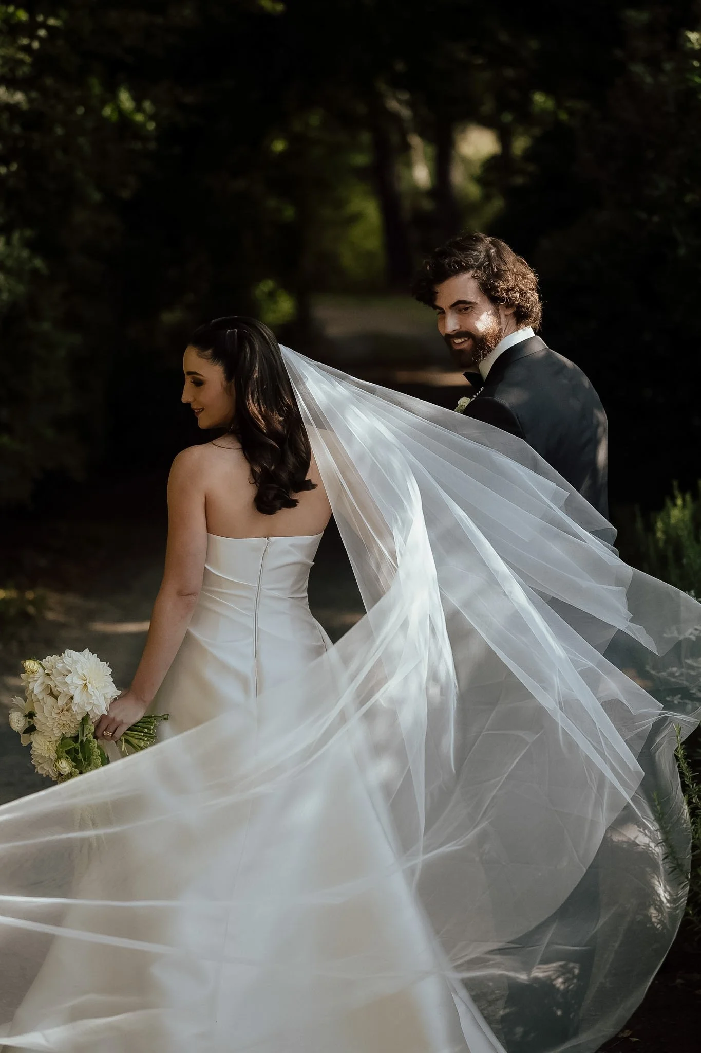Braidwood Wedding Photographer. A bride and groom standing outdoors surrounded by trees on a sunny day, with the bride wearing a white strapless wedding gown and holding a bouquet of white flowers, and the groom in a black tuxedo with a bow tie, both