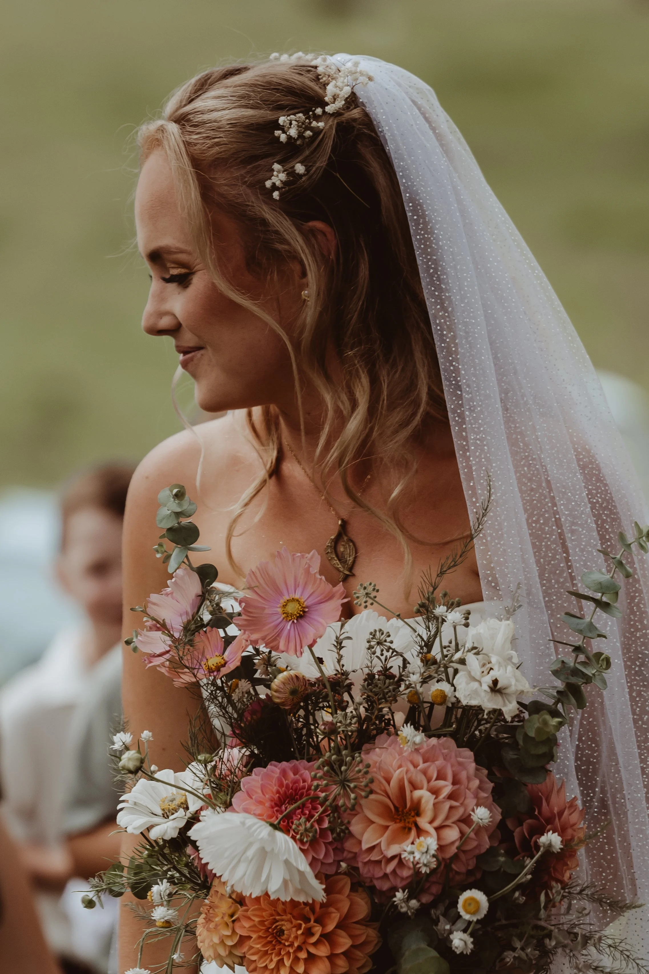 Braidwood Wedding Photographer. A woman, likely a bride, with long wavy hair and wearing a sheer veil is holding a large bouquet of flowers. She is smiling softly and has makeup on with arched eyebrows. The bouquet includes pink, white, and orange fl