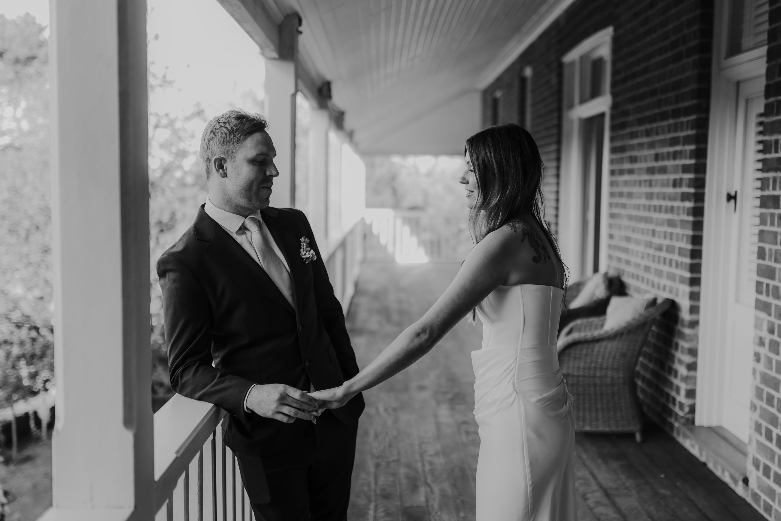Braidwood Wedding Photographer. A black and white photo of a couple holding hands on a porch, facing each other, with the woman wearing a wedding dress and the man in a suit.