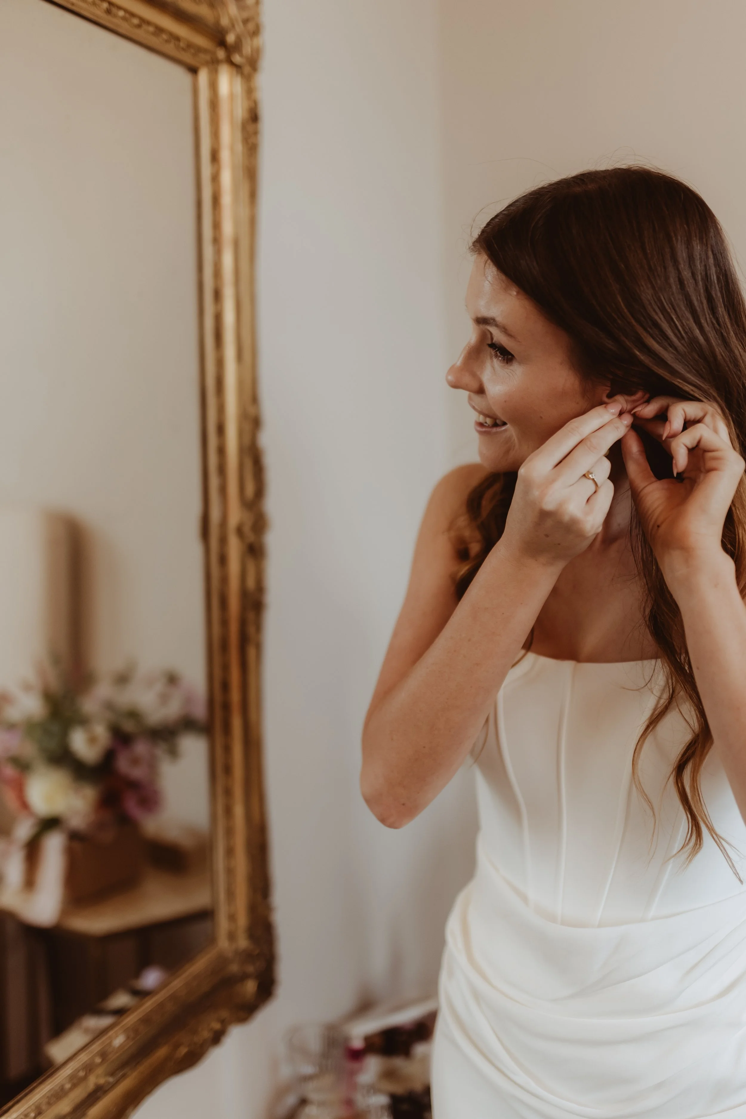 Braidwood Wedding Photographer. A woman with long brown hair putting on earrings, standing in front of a large, ornate gold-framed mirror in a room with neutral walls.