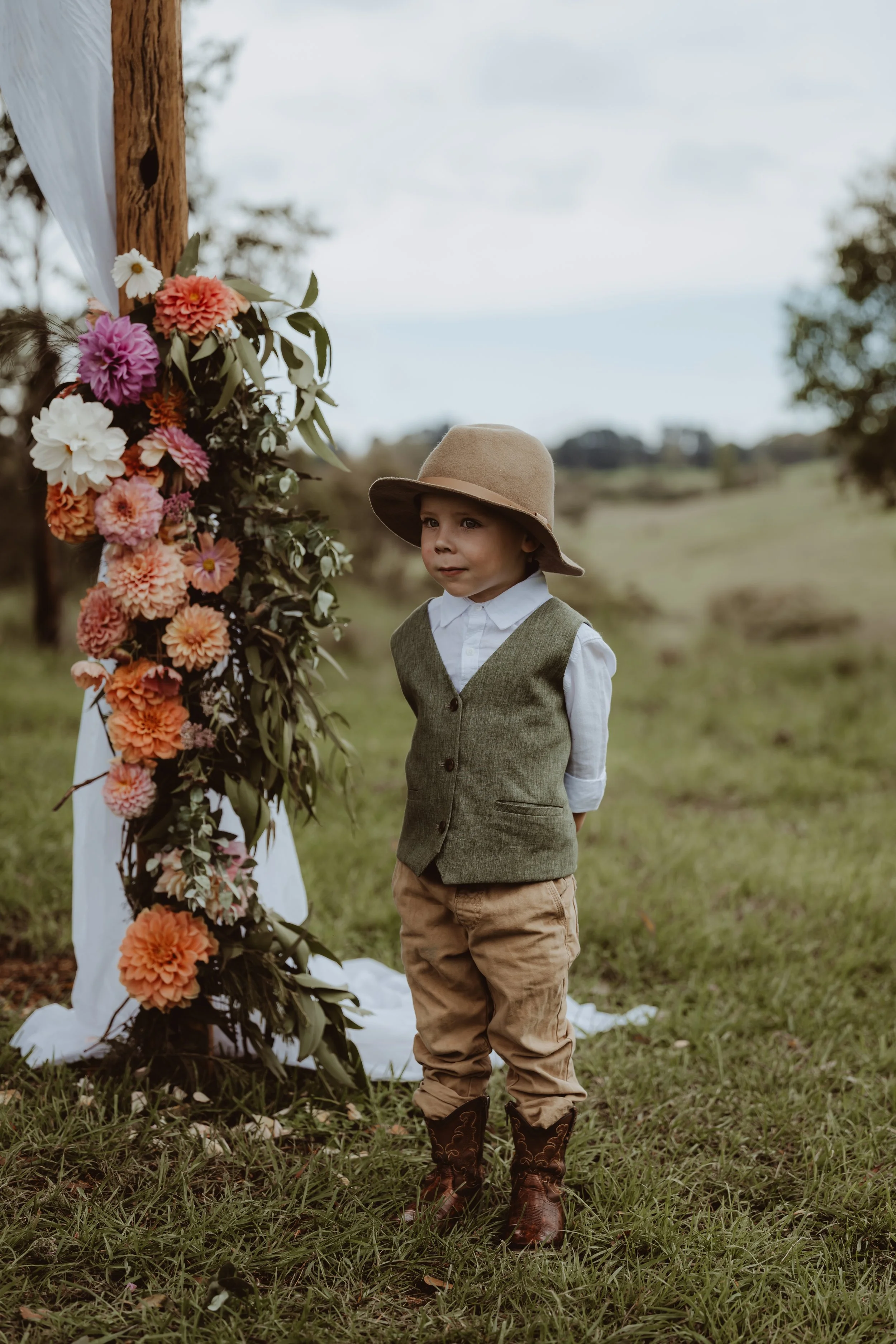 Braidwood Wedding Photographer. A young boy wearing a beige hat, white shirt, brown vest, and tan pants stands outdoors in a grassy field with a decorated floral arch behind him. The arch is adorned with pink, peach, and white flowers, and green leav