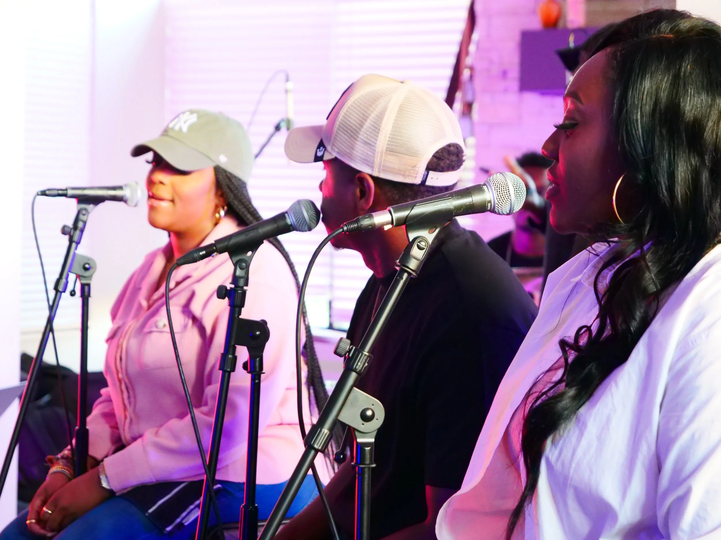 Three women and one man sitting in a row with microphones, possibly singing or speaking, in a colorful indoor setting.