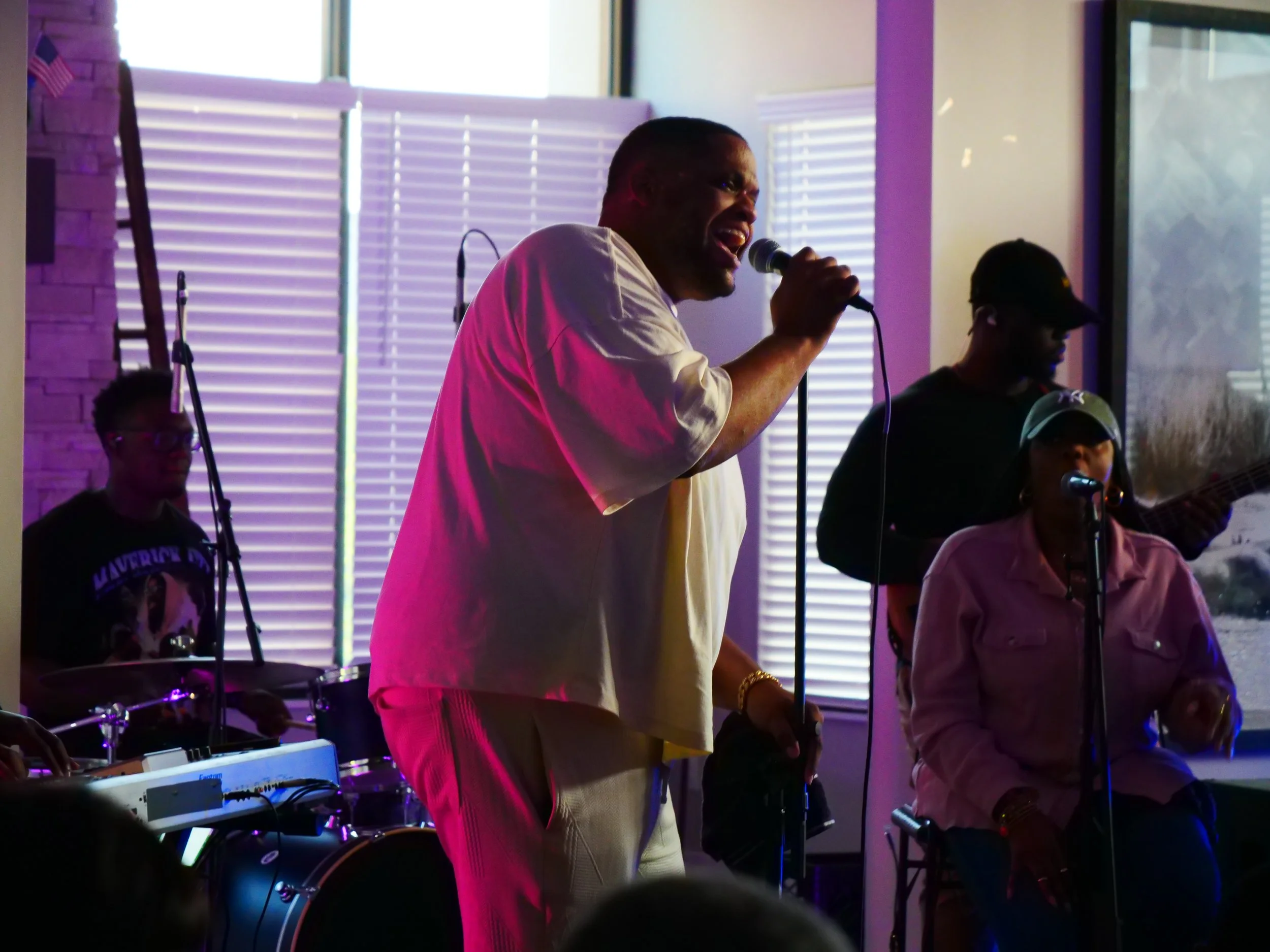 A man singing into a microphone with a band playing behind him, including a drummer and a woman with a microphone and a guitarist, in a dimly lit indoor setting.
