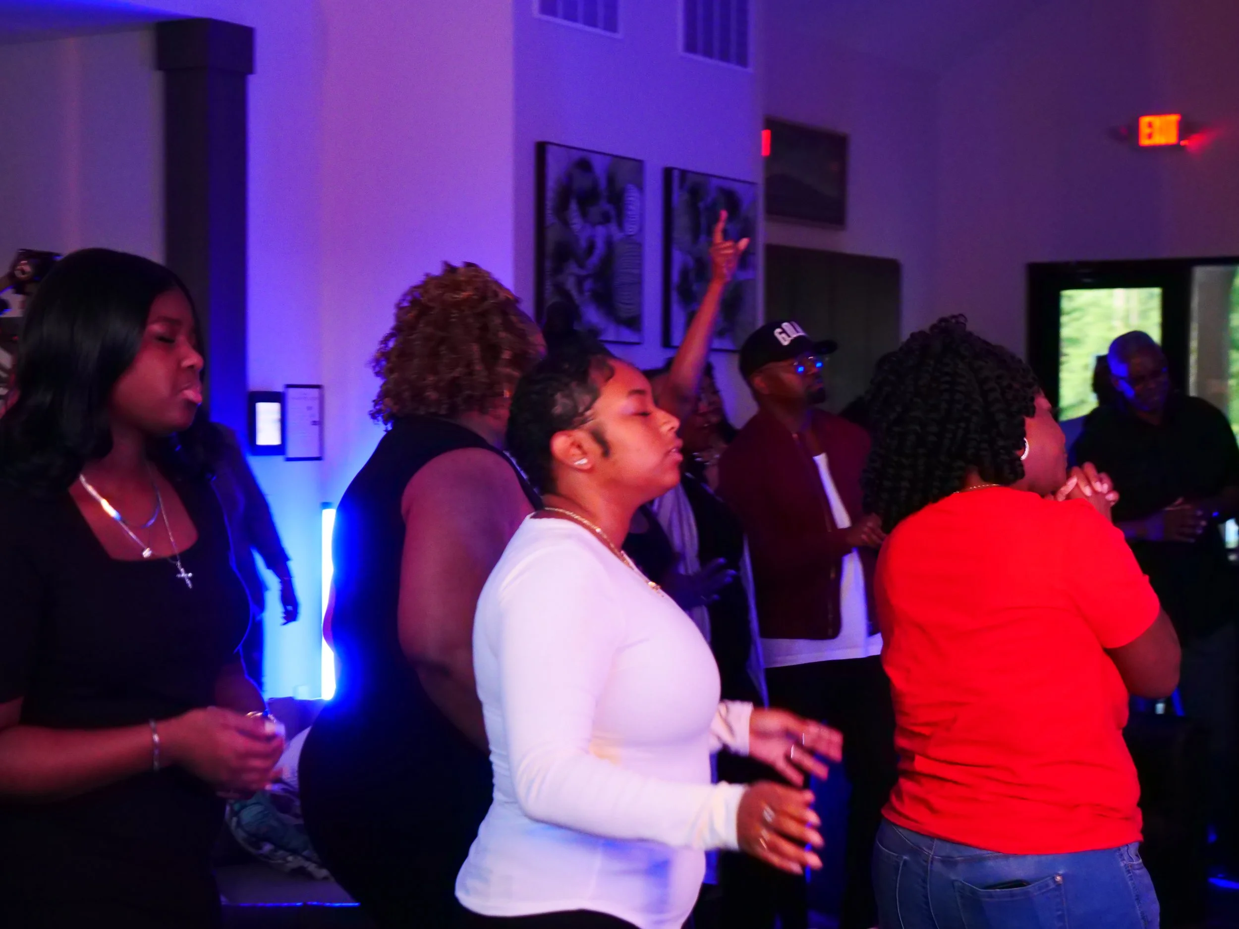 Group of people at a party or gathering, some with eyes closed, dancing or enjoying music in a dimly lit room with colorful lighting.